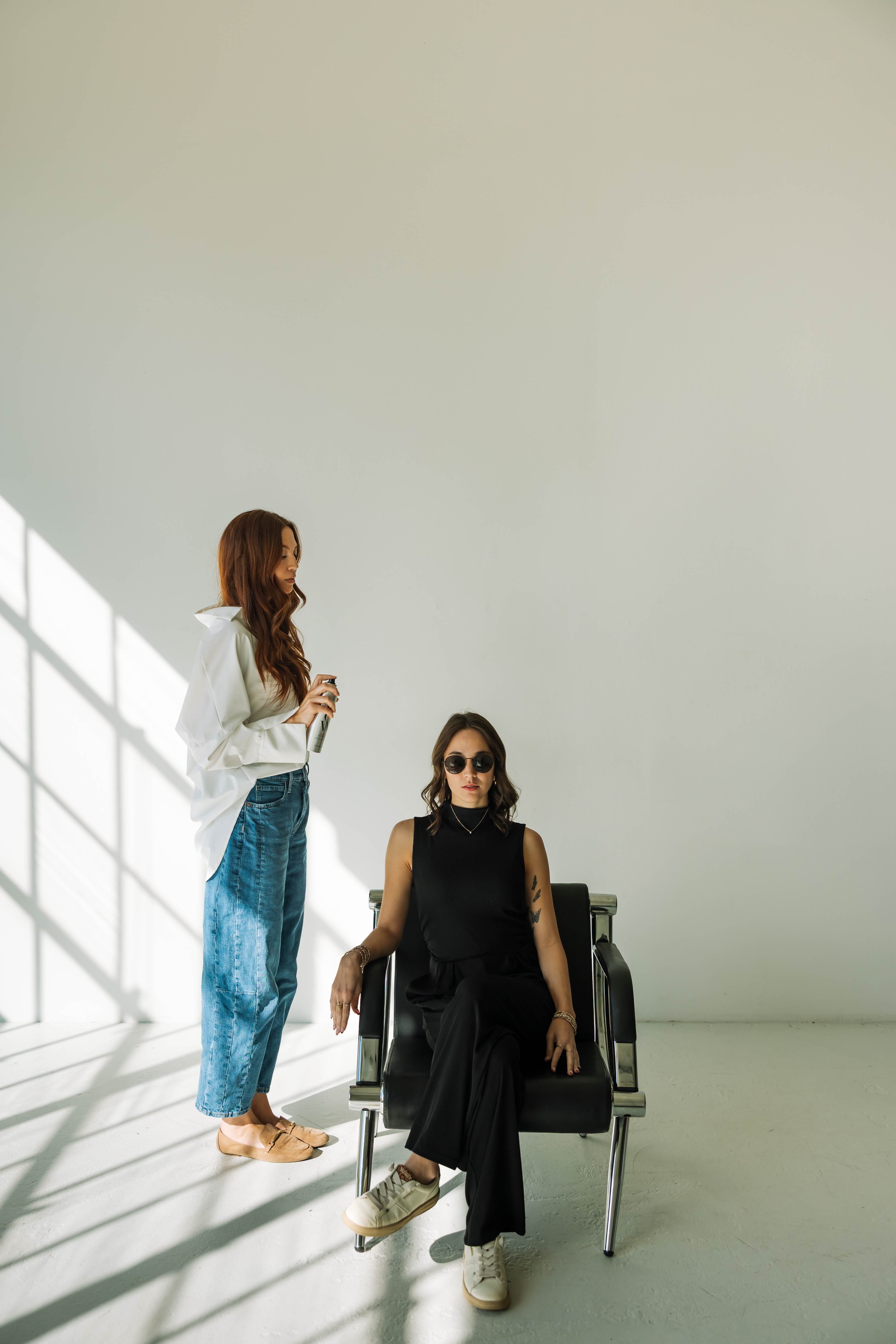 Two women in a minimalist room with sunlight and shadows. One woman with long brown hair in a white shirt and jeans stands holding a spray bottle, while the other woman with dark hair, sunglasses, and tattoos sits in a black chair wearing a black sleeveless top and pants.