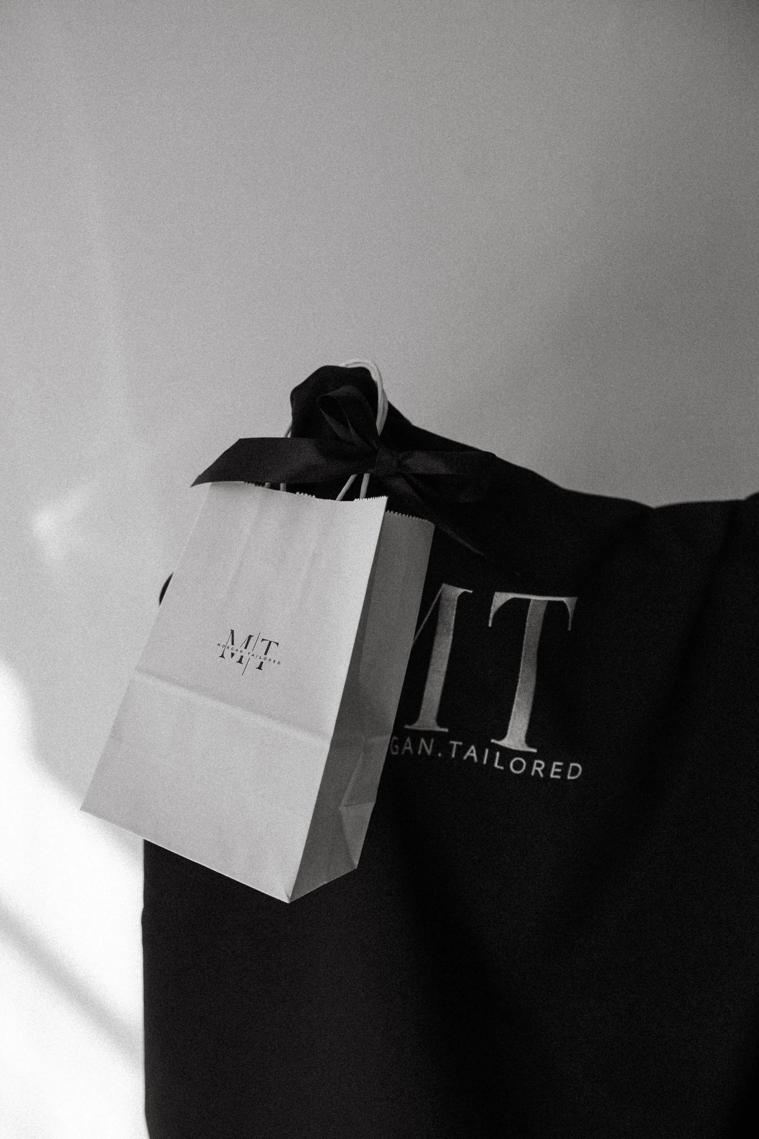 A person holding a shopping bag in a black sweatshirt with the words "TIGER. TAILORED" printed on it. The bag has the brand logo and is tied with a ribbon.