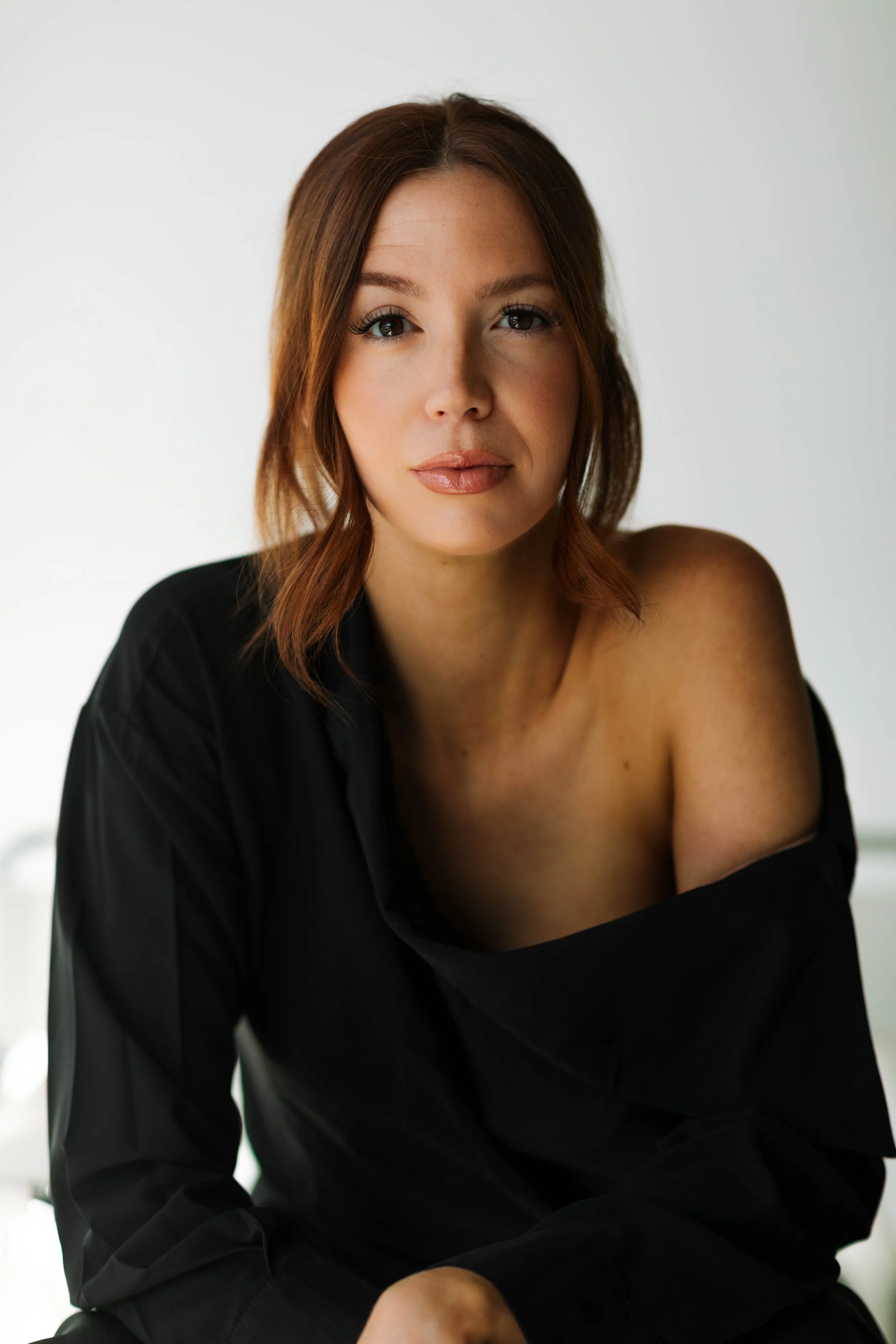 A woman with shoulder-length brown hair, wearing a black off-shoulder top, sitting against a neutral background, looking directly at the camera.