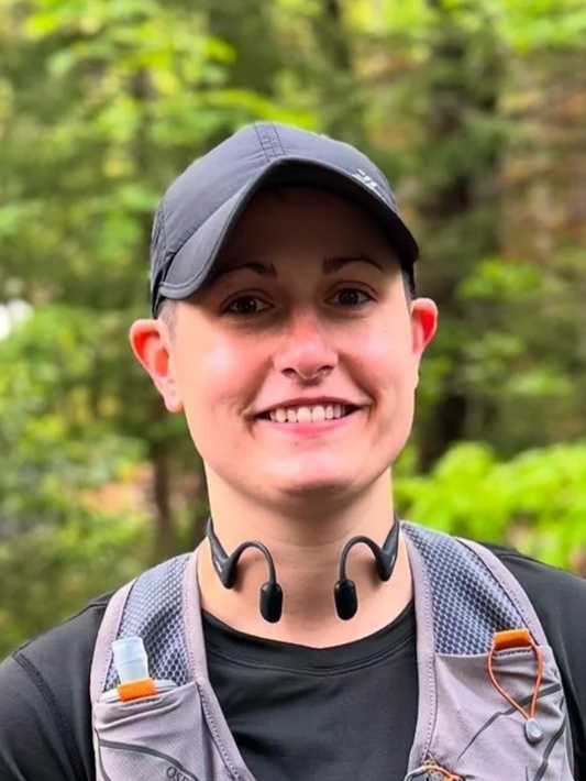 Smiling woman wearing a gray cap, black shirt, and a gray hydration backpack outdoors with green trees in the background. Wayfinder Running Co