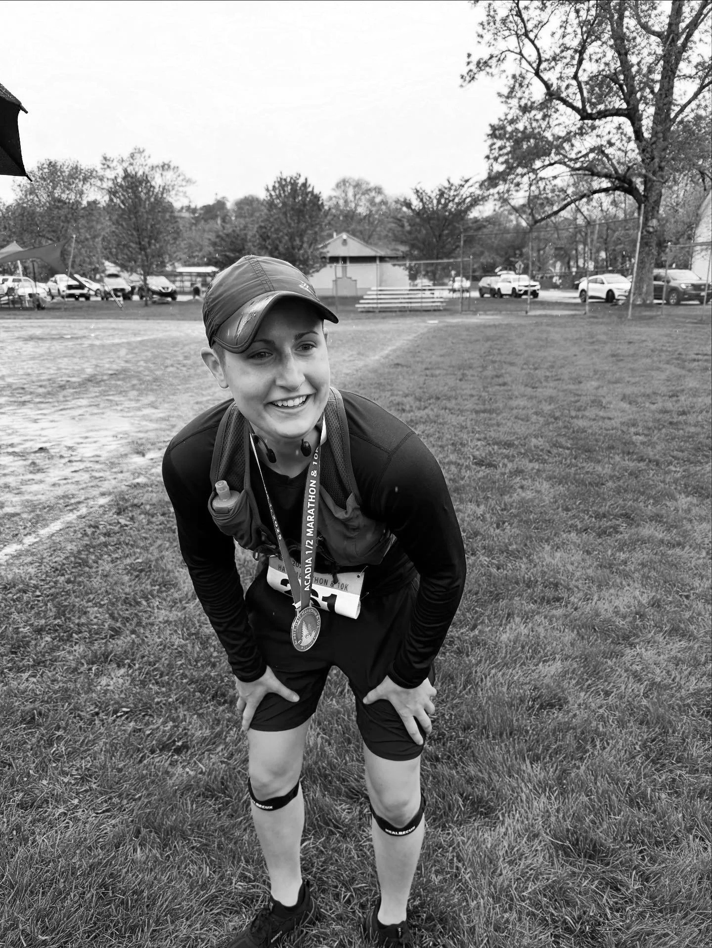 A woman in athletic gear with a medal around her neck, smiling and leaning forward on a grassy field after a race. Wayfinder Running Co