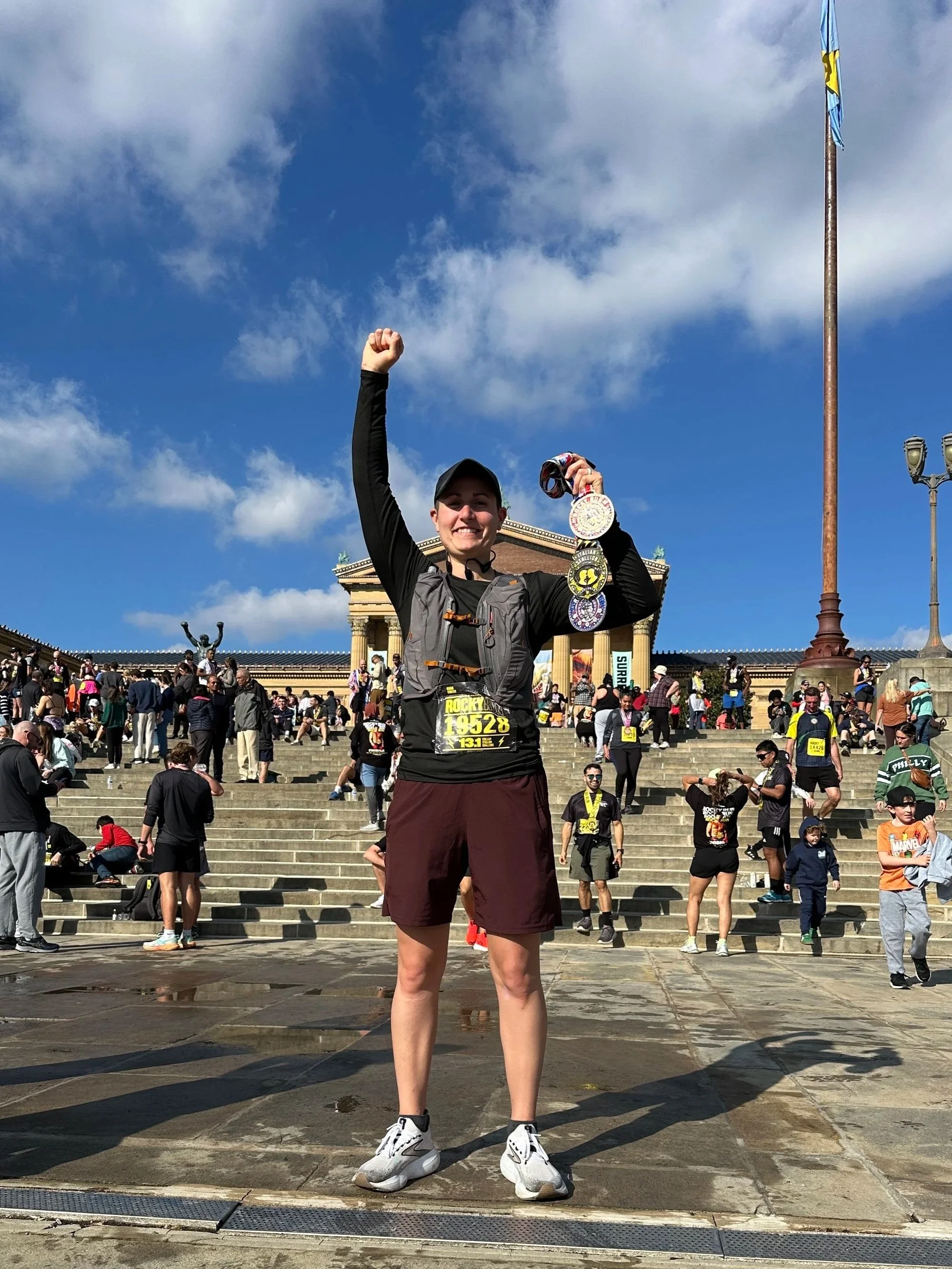 Coach Lauren,RRCA Certified Running Coach and ISSA Certified Nutrition Coach, standing on the steps of the Philadelphia Museum of Art, smiling, holding medals, wearing a race bib, with a large crowd of people behind her.