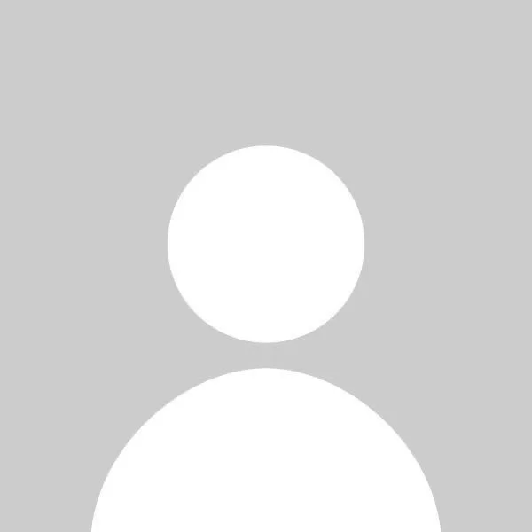 A man sitting on a beige bench against a light-colored wall, looking to his left, wearing a blue short-sleeve sweater and beige pants, with his hands resting on the bench.