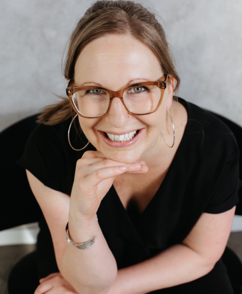 A smiling 45-year-old female, wearing brown framed glasses and a black top, leaning her chin on her hand.