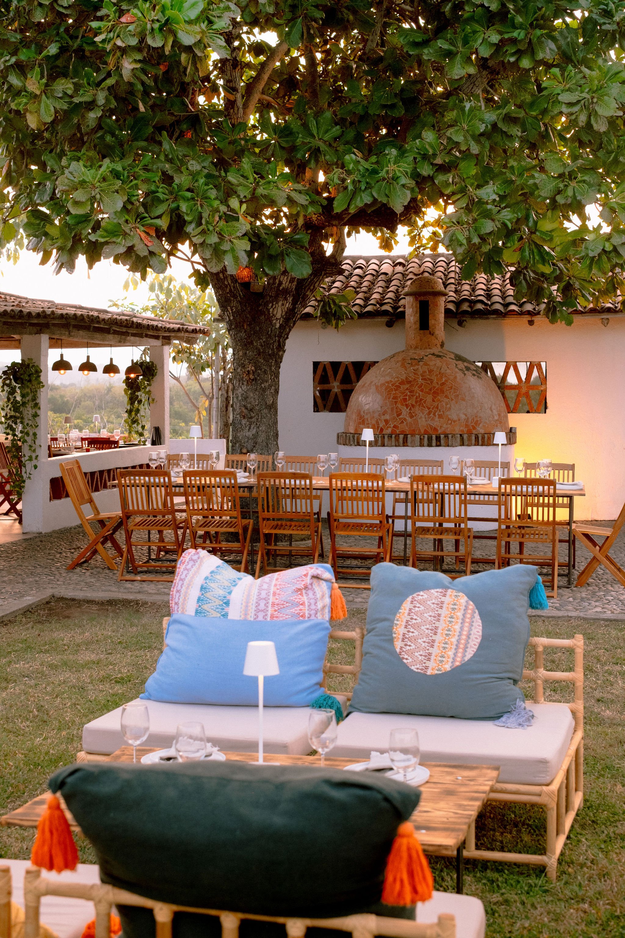 Una mesa de comedor al aire libre con muchas sillas de madera, ubicada frente a una chimenea de barro, bajo un árbol grande con follaje verde, y decorada con copas y utensilios para comer