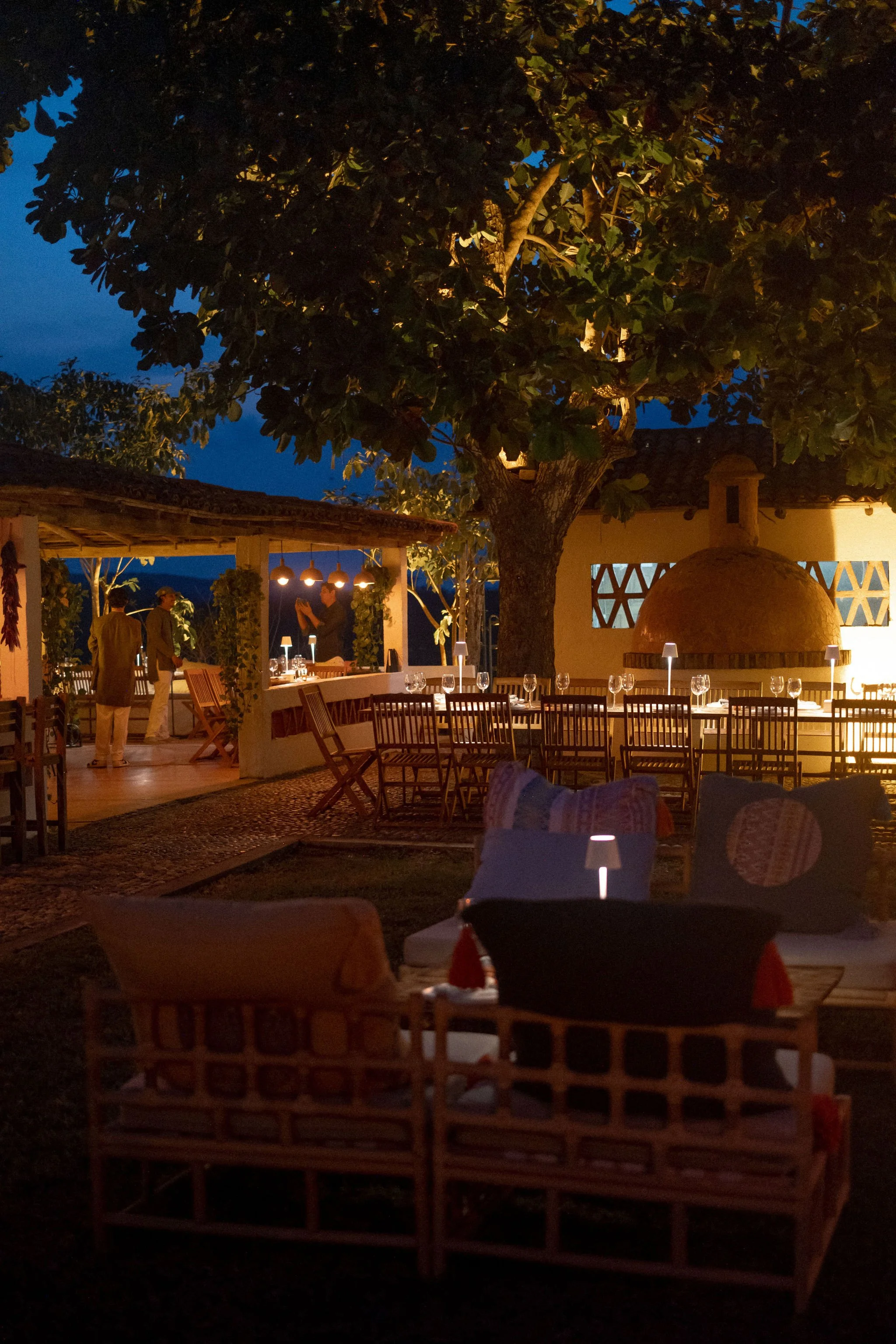 Escena de una cena al aire libre en POLO CAREYES un jardín al atardecer con mesas, sillas y un horno de pizza de ladrillo.