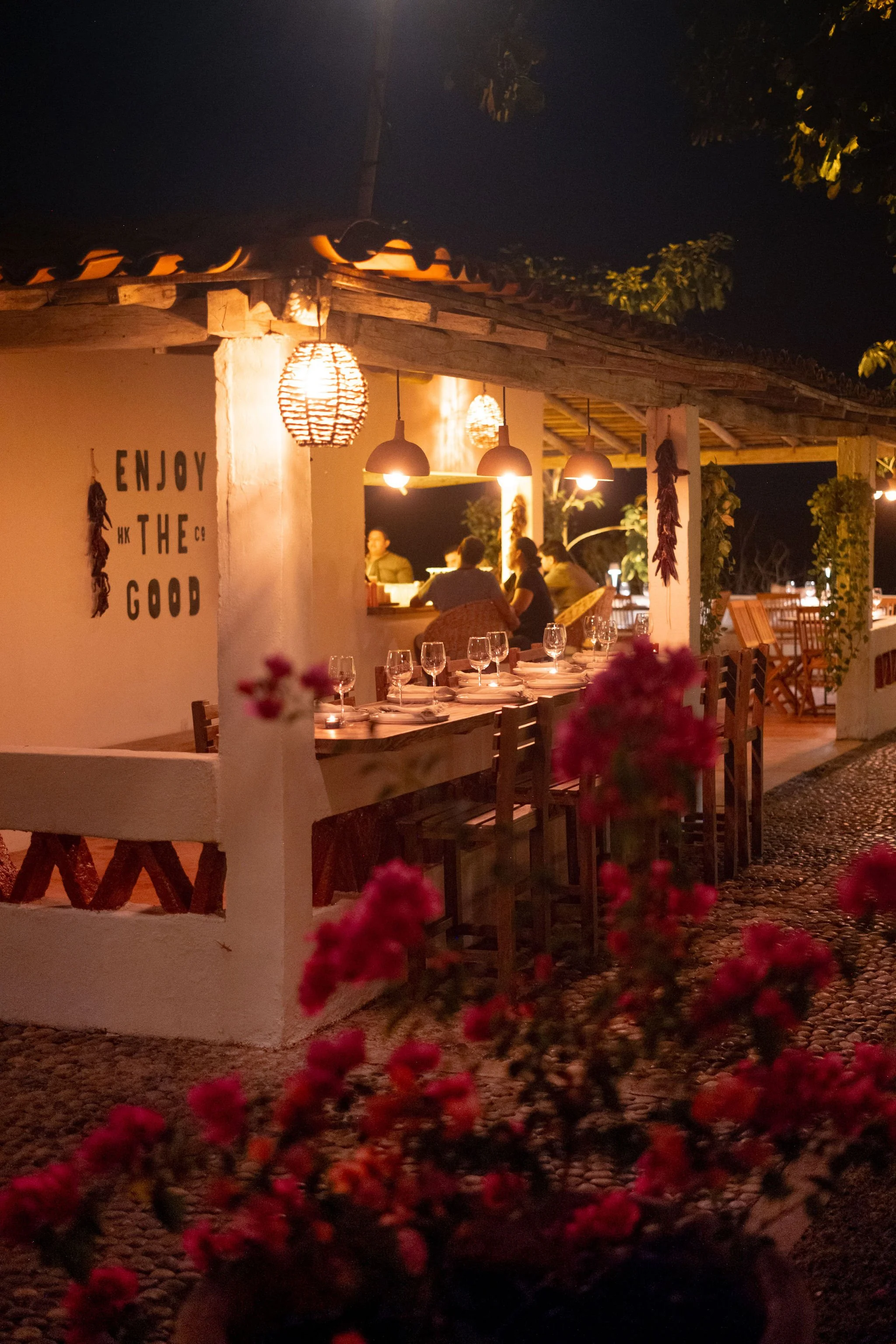 Una escena de una terraza de restaurante malabar careyes con mesas decoradas para cenar, en la noche, con luces colgantes y algunas flores en primer plano, y un grupo de personas en la barra de atención al cliente en el fondo.