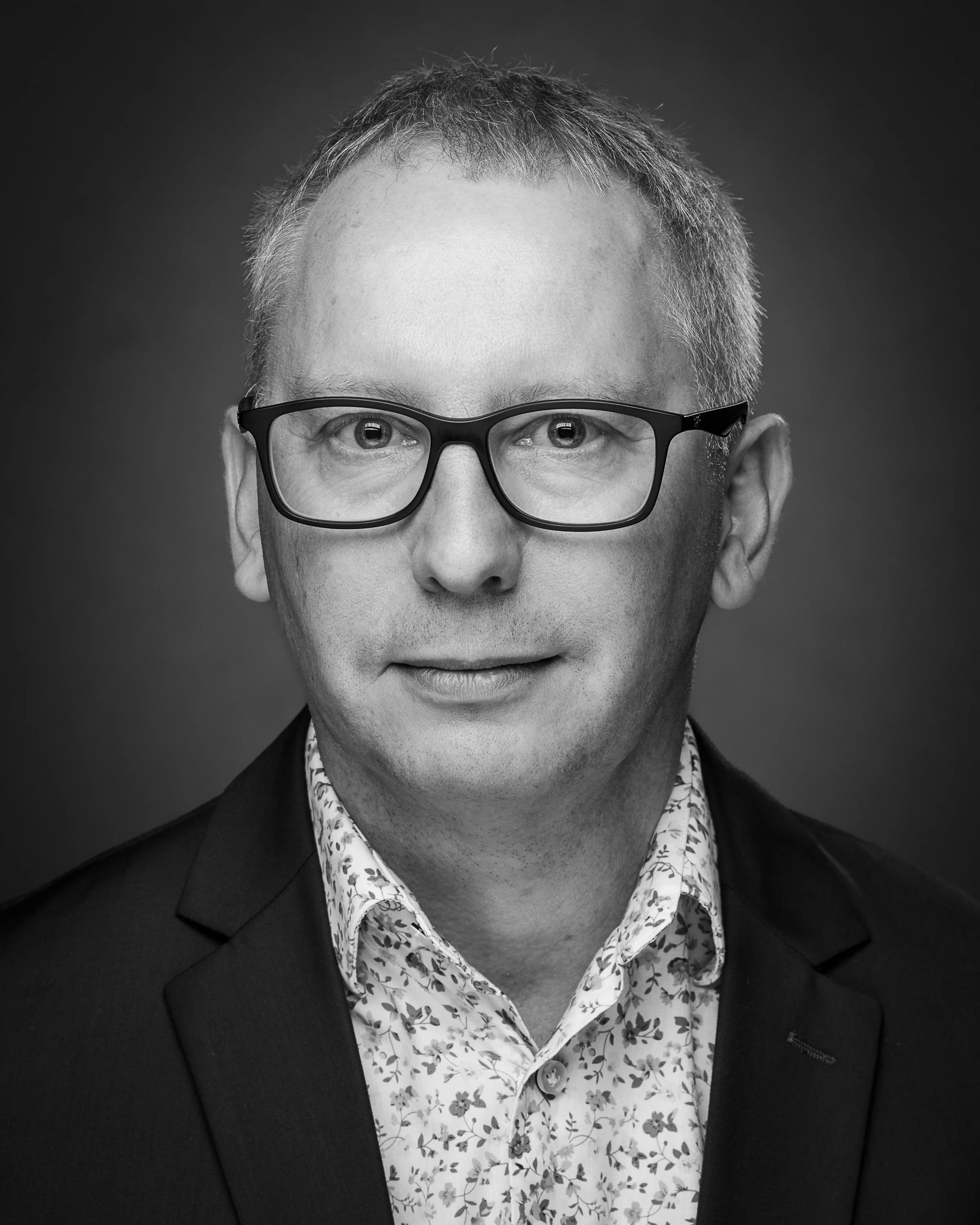 Black and white portrait of a man wearing glasses, a floral patterned shirt, and a blazer, looking directly at the camera.