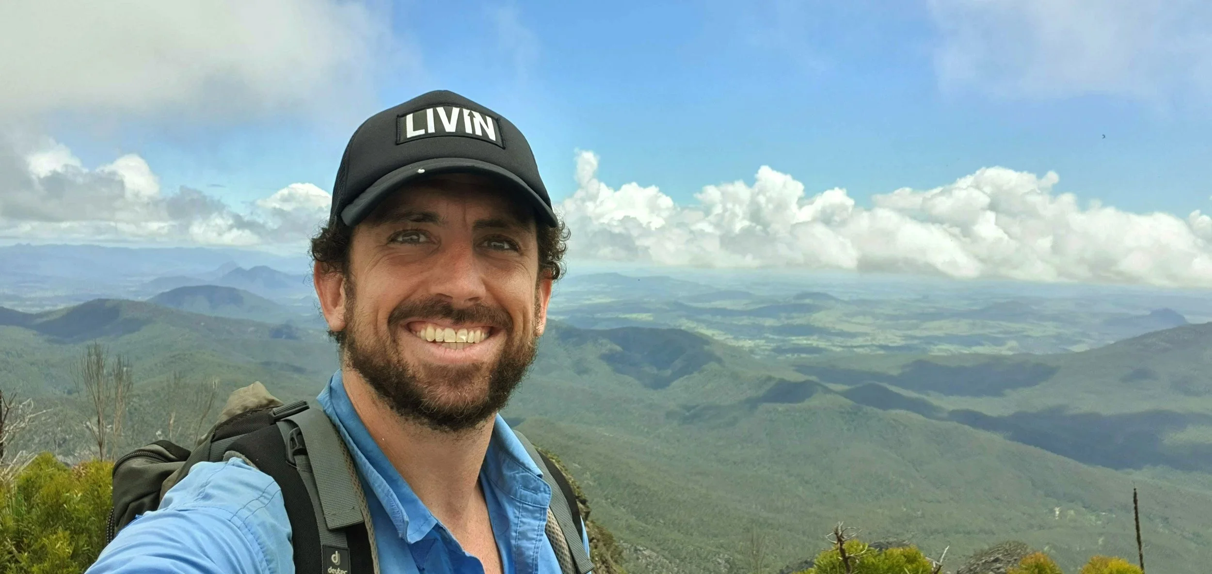 A man smiling outdoors in a mountainous area with green hills and a partly cloudy sky.