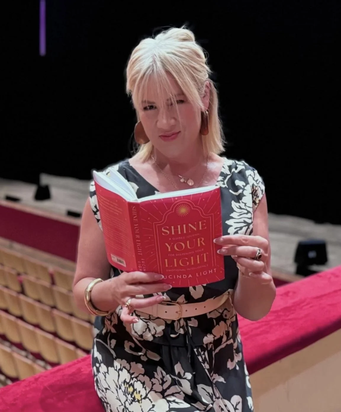 A woman with blonde hair reading a red book titled 'Shine Your Light' in a theater or auditorium with empty seats and a red velvet railing.