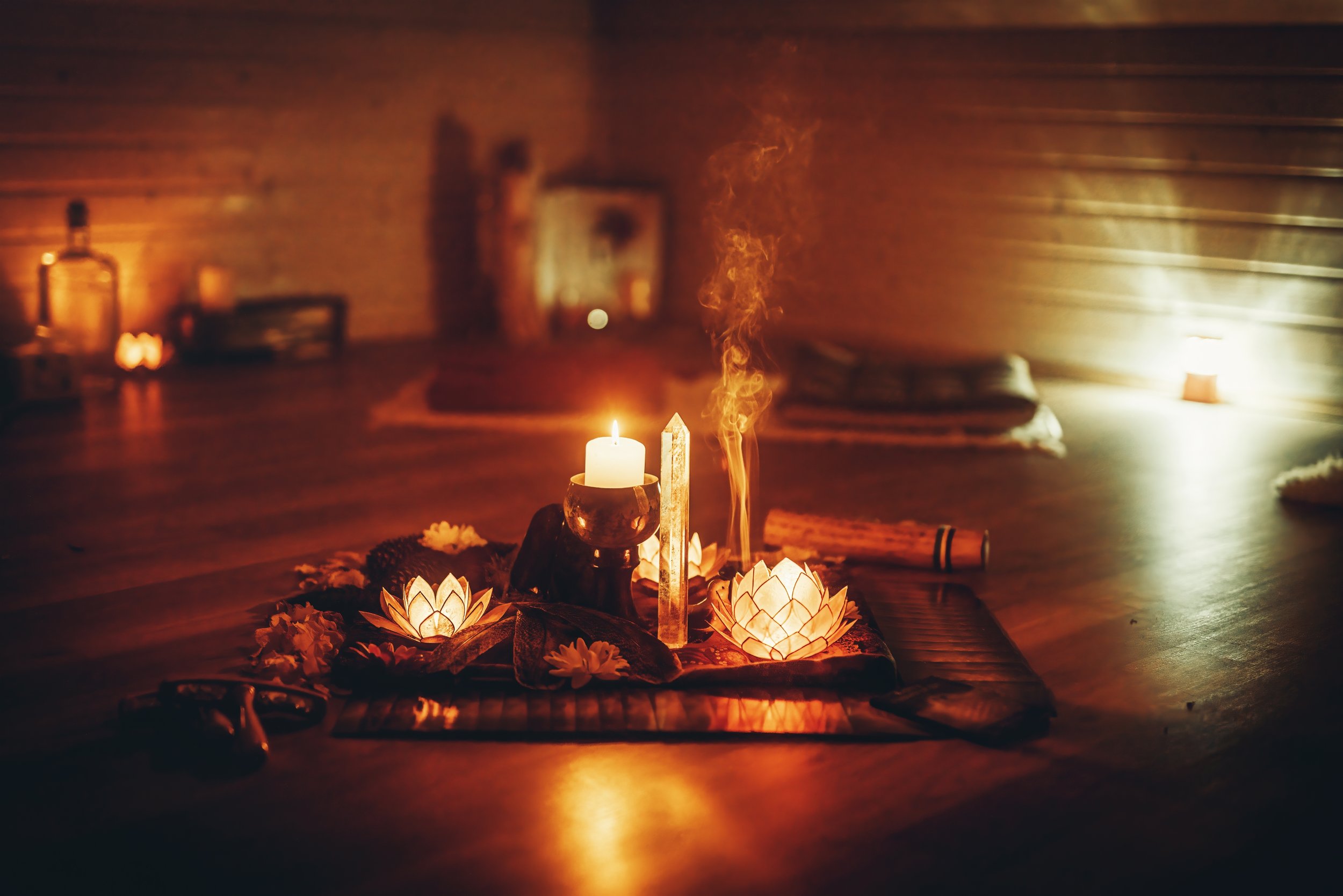 A meditation or altar setup on the wooden floor of a dimly lit room with candles, lotus-shaped lights, crystals, and incense, creating a warm, peaceful atmosphere.