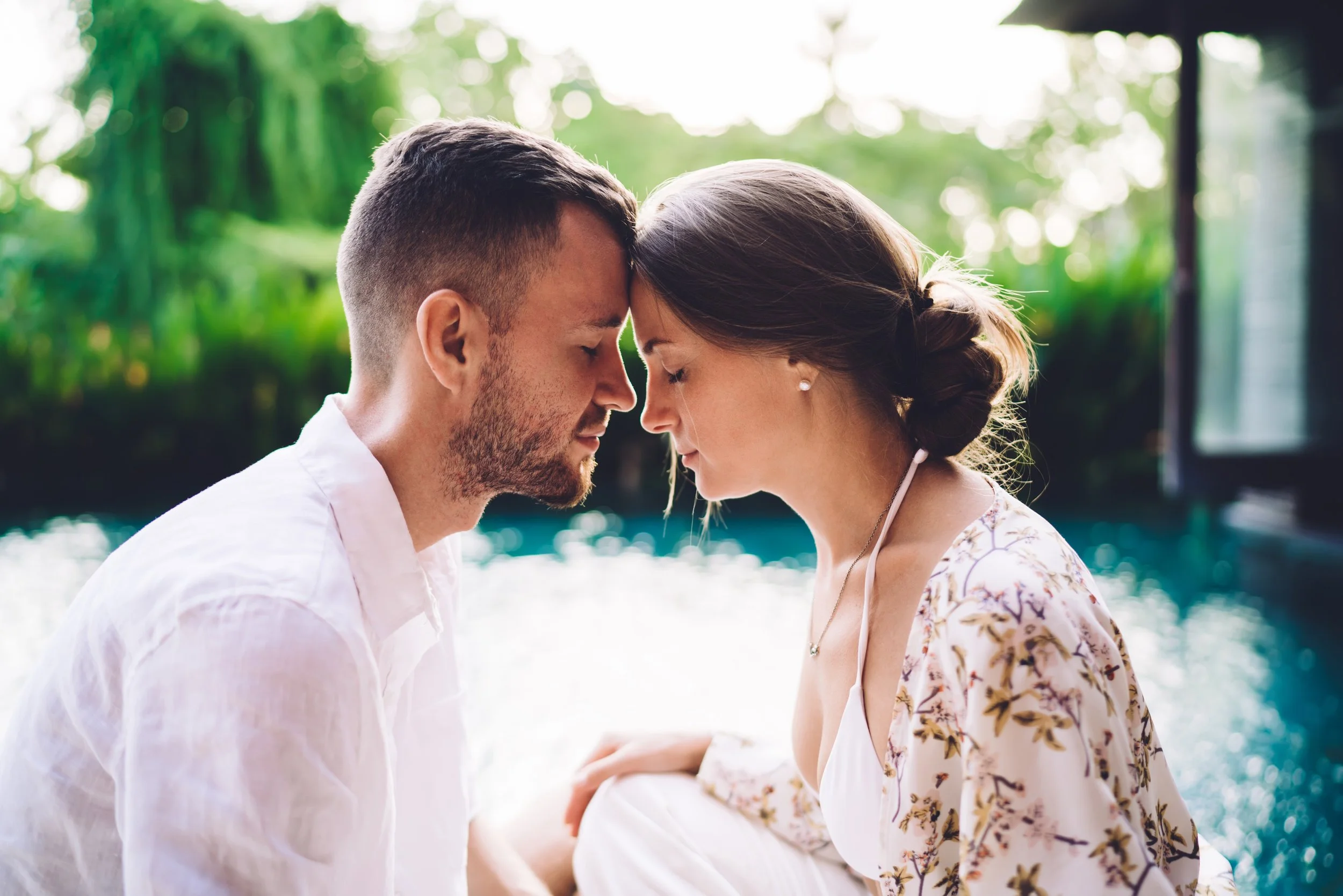 A couple sitting close together with foreheads touching near a pool, with greenery in the background.
