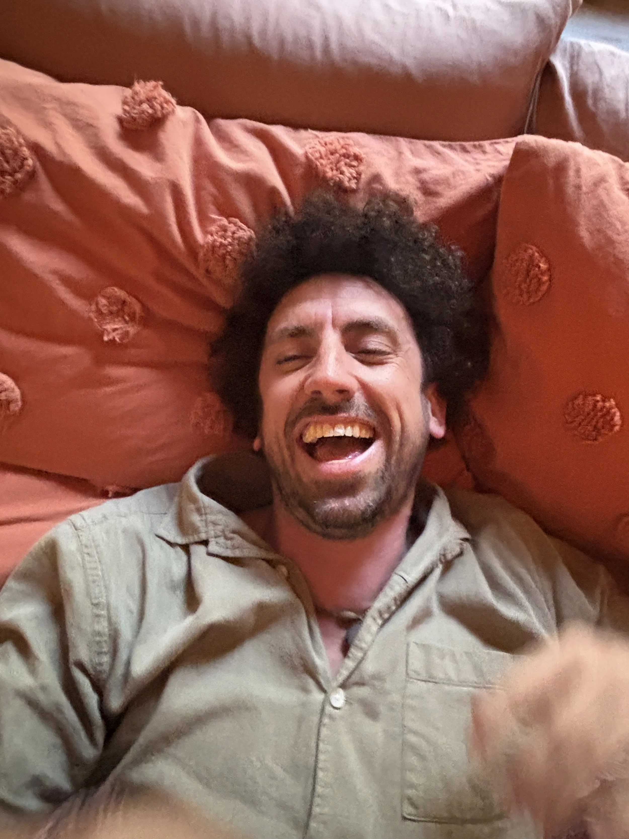A man with curly black hair is lying on a reddish-orange pillow and pillowcases, smiling or laughing with his eyes partially closed.