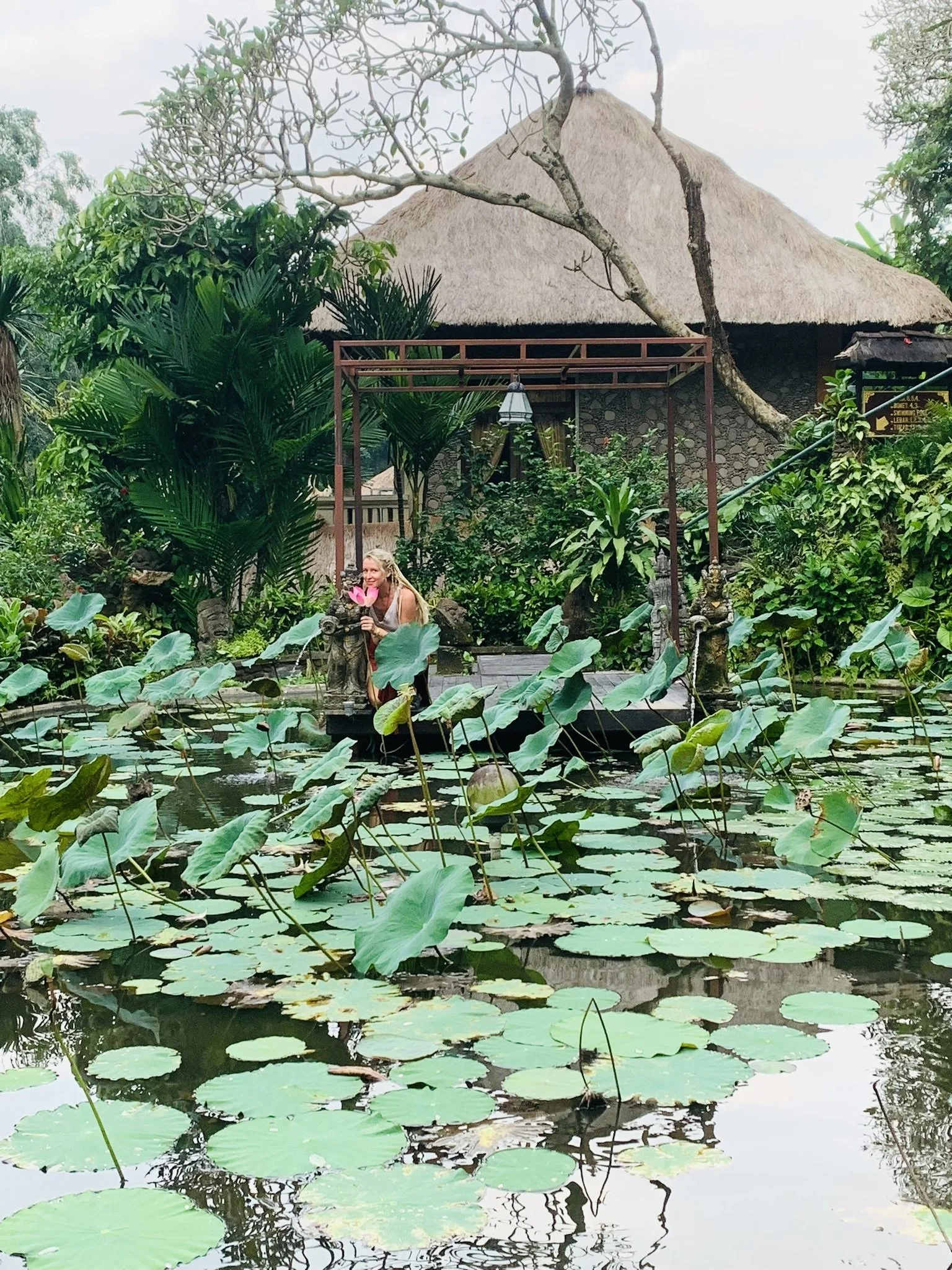 A woman with blonde hair holding a pink lotus flower at the edge of a pond filled with green lily pads and lotus plants, surrounded by lush tropical foliage, with thatched-roof building in the background.
