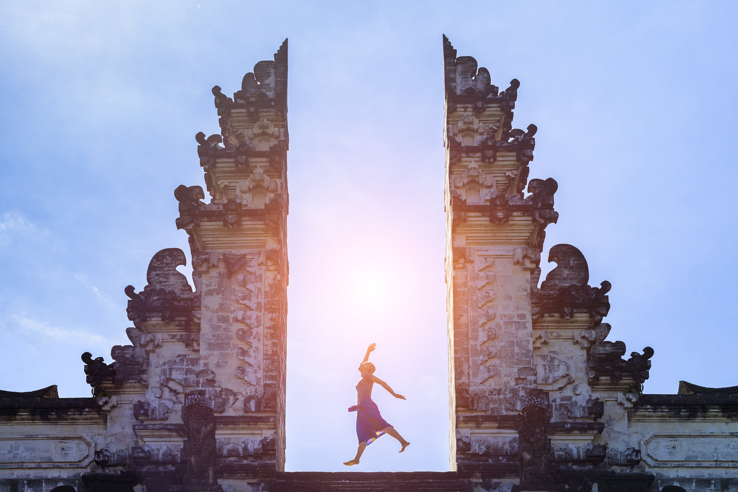 A person jumping between two ornate stone temple towers with a bright sun in the background and a clear blue sky.