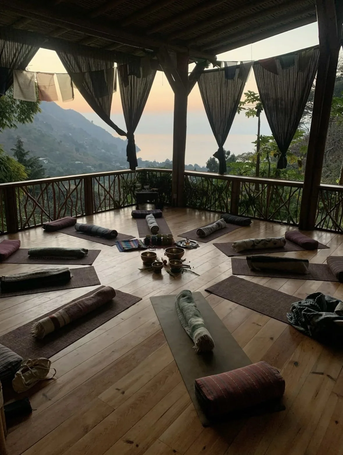 A circular yoga or meditation space on a wooden deck with rolled mats, cushions, and bowls, overlooking a valley and mountains at sunset.