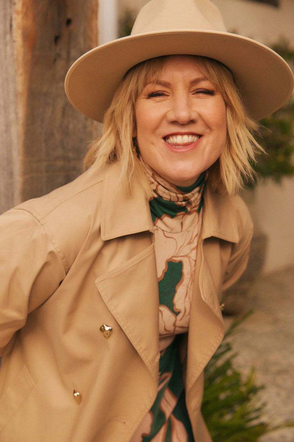 A woman smiling with short blonde hair, wearing a tan hat, tan trench coat, and a patterned scarf.