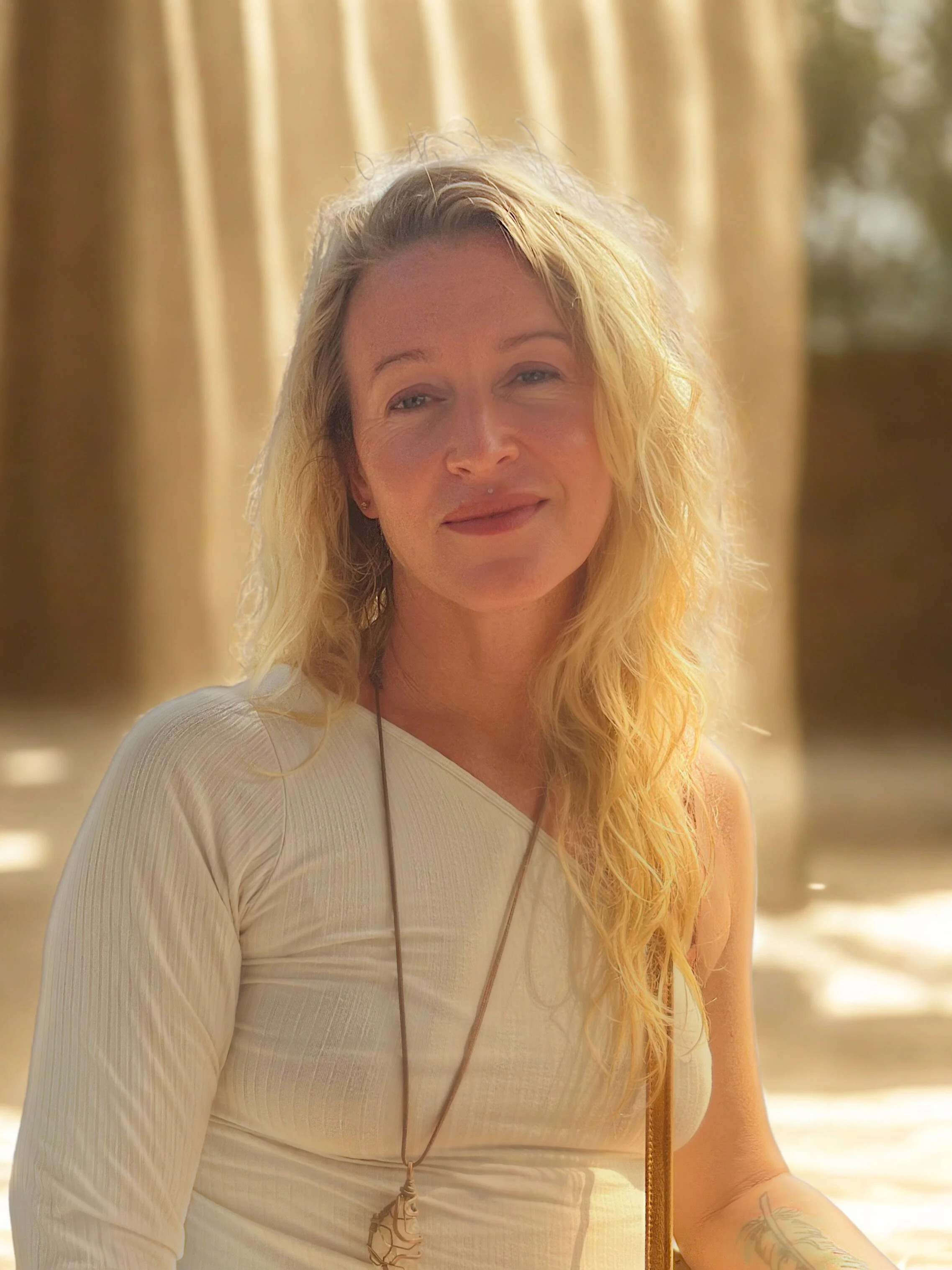 A woman with blonde wavy hair smiling softly, wearing a cream-colored long-sleeve top and a long necklace, standing outdoors with beige curtains and trees in the background, bathed in warm natural sunlight.