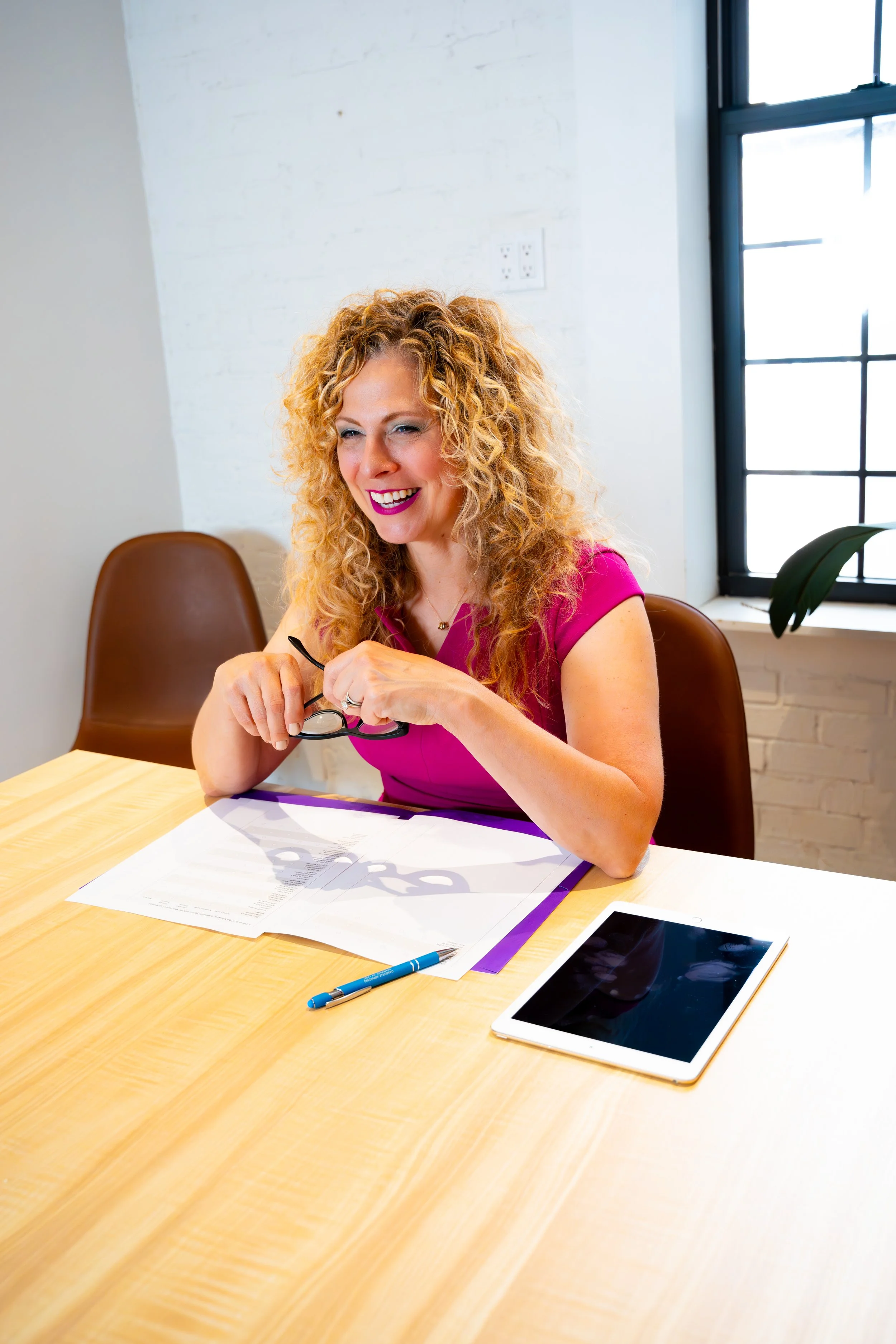 A woman with curly red hair, wearing a pink top, sitting at a light wooden table with papers and a tablet, smiling and holding glasses, in a bright room with a large window and white brick wall.