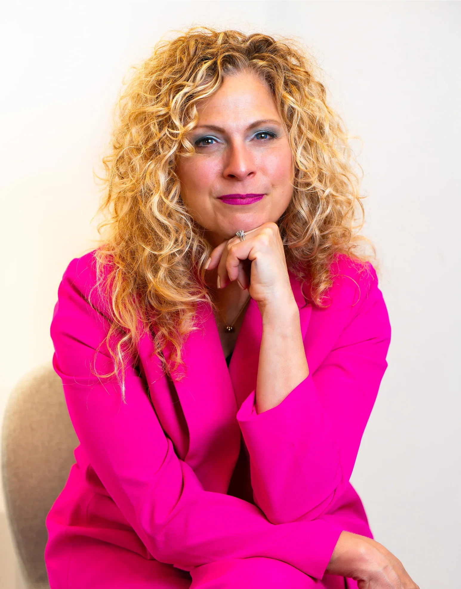 Portrait of a woman with curly blonde hair wearing a bright pink blazer, sitting with her hand resting under her chin, against a plain light background.