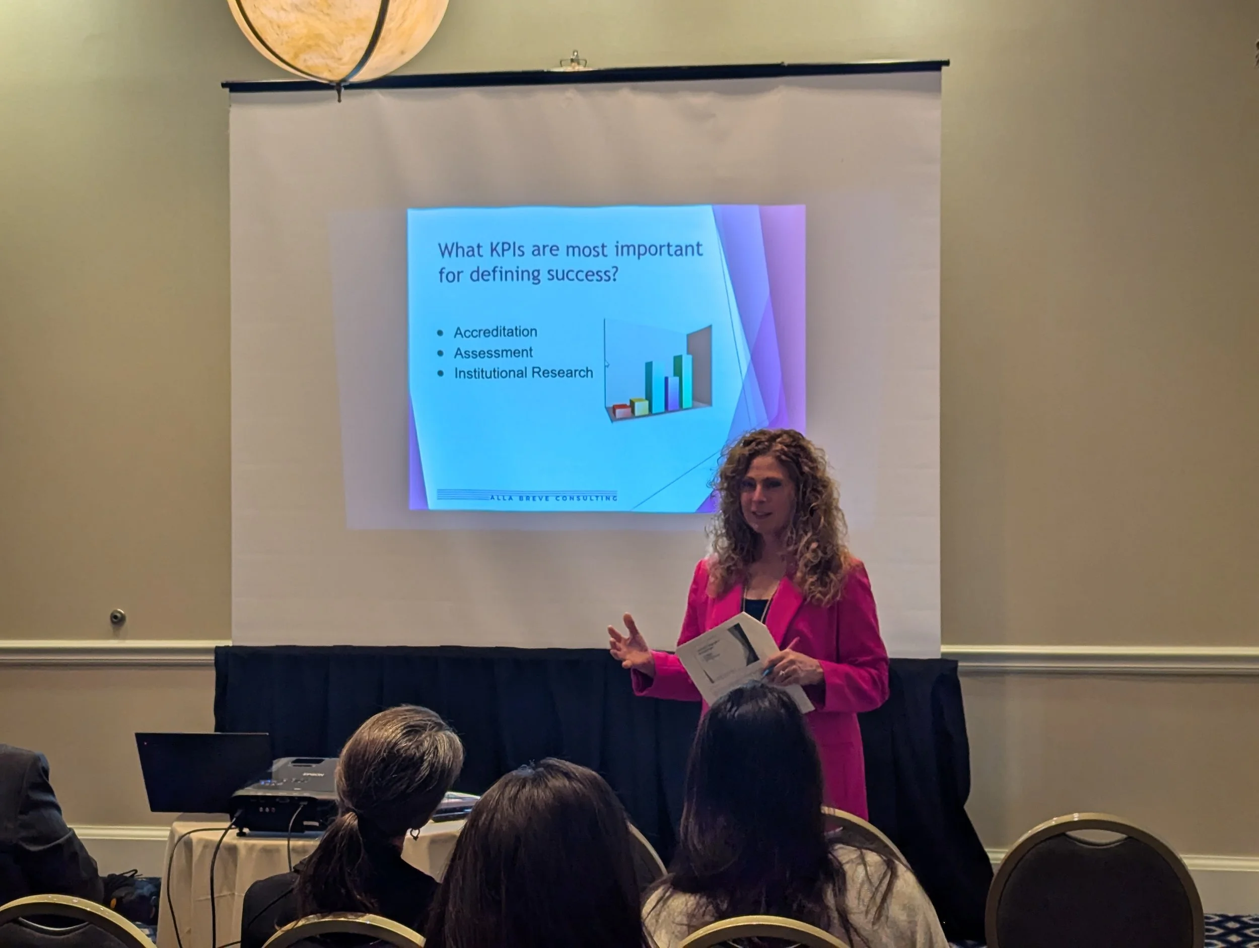 A woman with curly hair, dressed in a pink blazer, is giving a presentation in front of a projected slide at a conference. The slide discusses KPIs related to success, including accreditation, assessment, and institutional research.