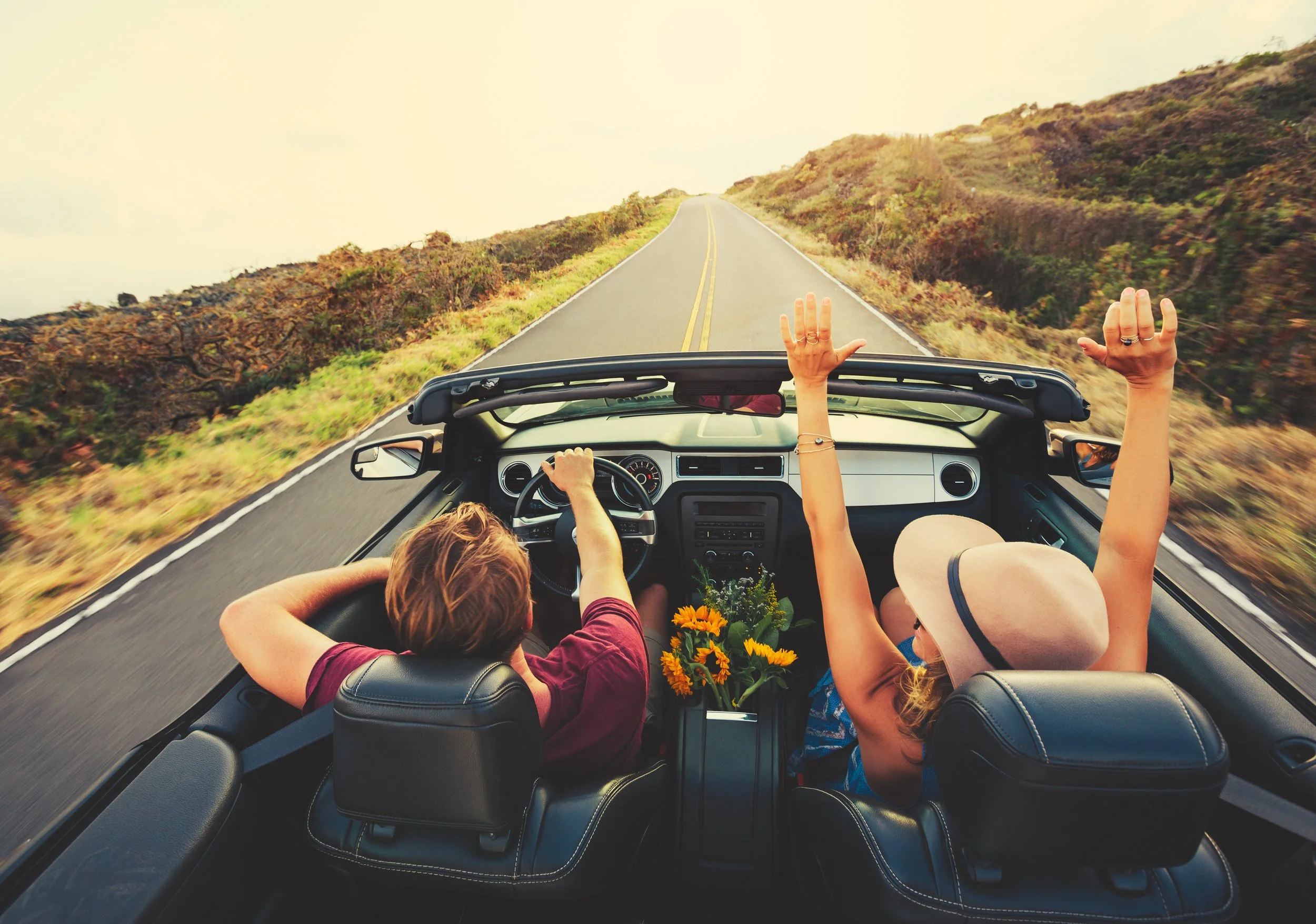 Two people in a convertible car driving on a scenic road through the countryside with hills and clear sky.