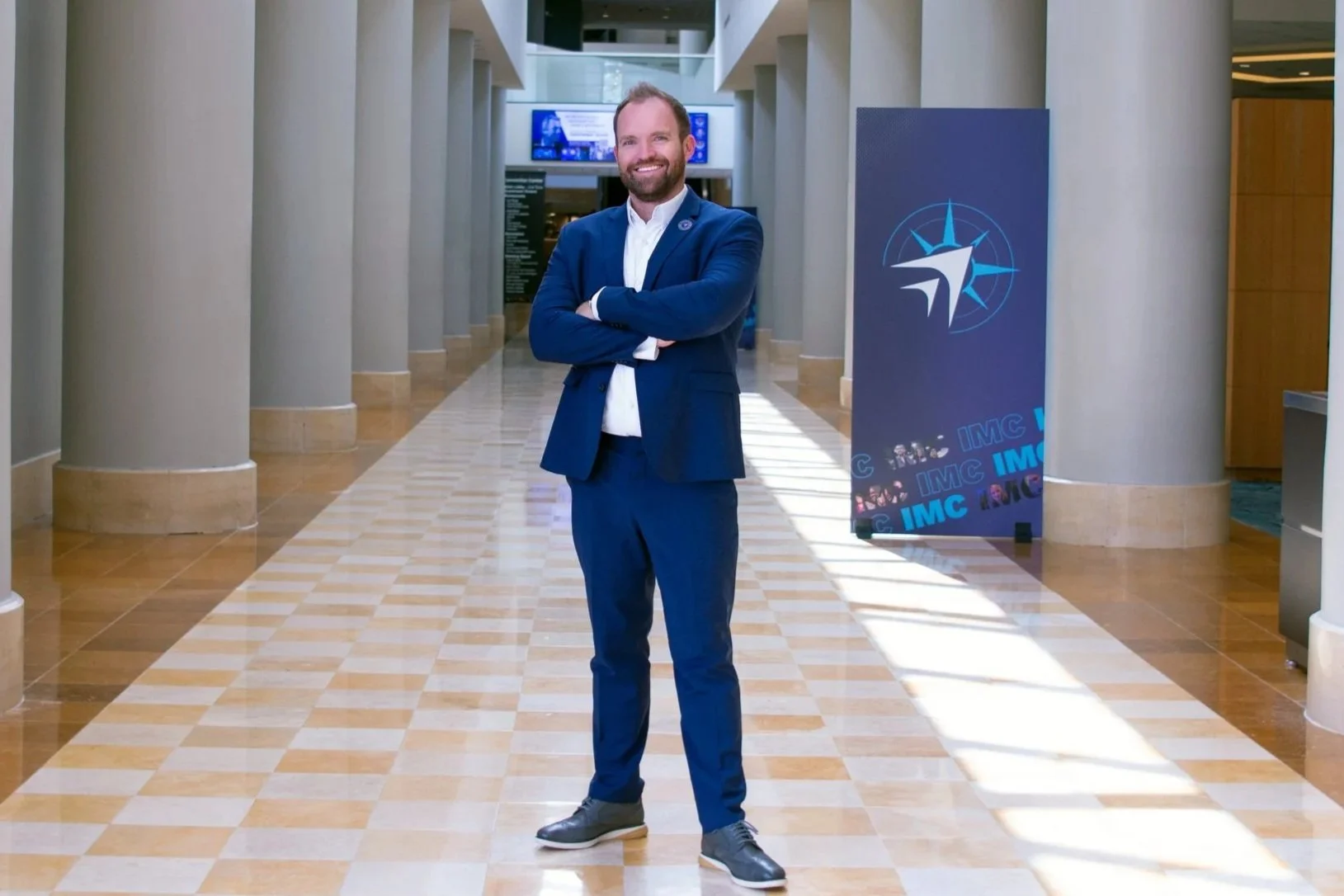 A man in a blue suit standing with arms crossed in a lobby area with large columns and a blue sign with a compass logo and the word 'IMC' repeated.