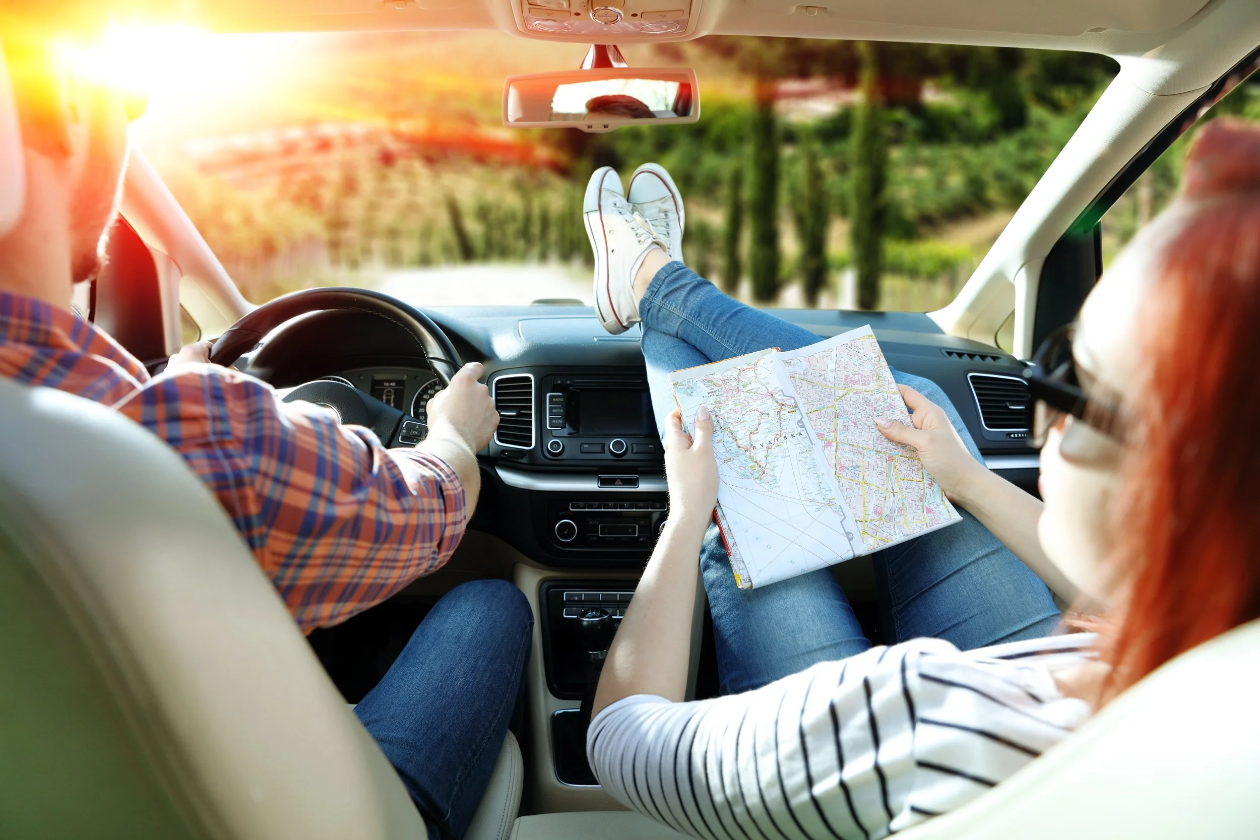 A couple on a road trip in a car, with the woman relaxing with her feet up on the dashboard, holding a map, while the man drives during sunset.