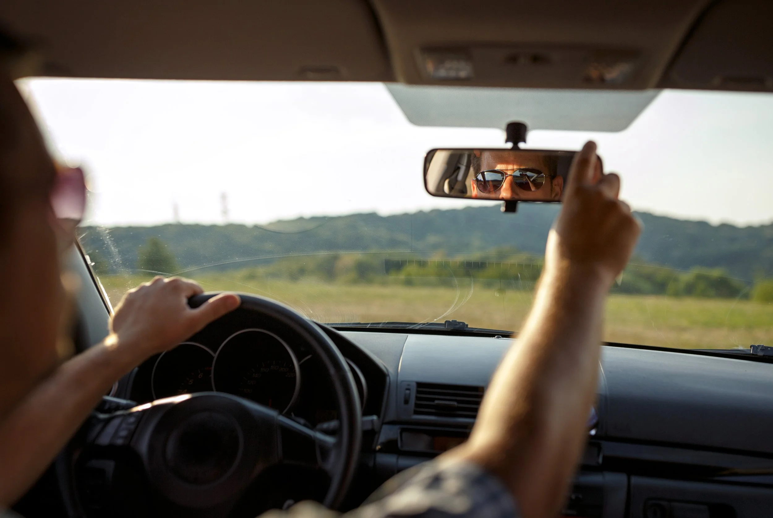 Inside view of a car showing a person adjusting the rearview mirror with sunglasses, with a landscape of green hills visible through the windshield.