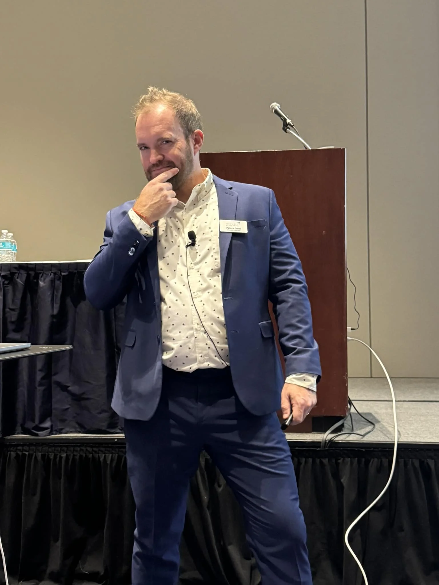 Man in blue suit with white shirt posing in front of podium with microphone, standing on stage at conference or event.