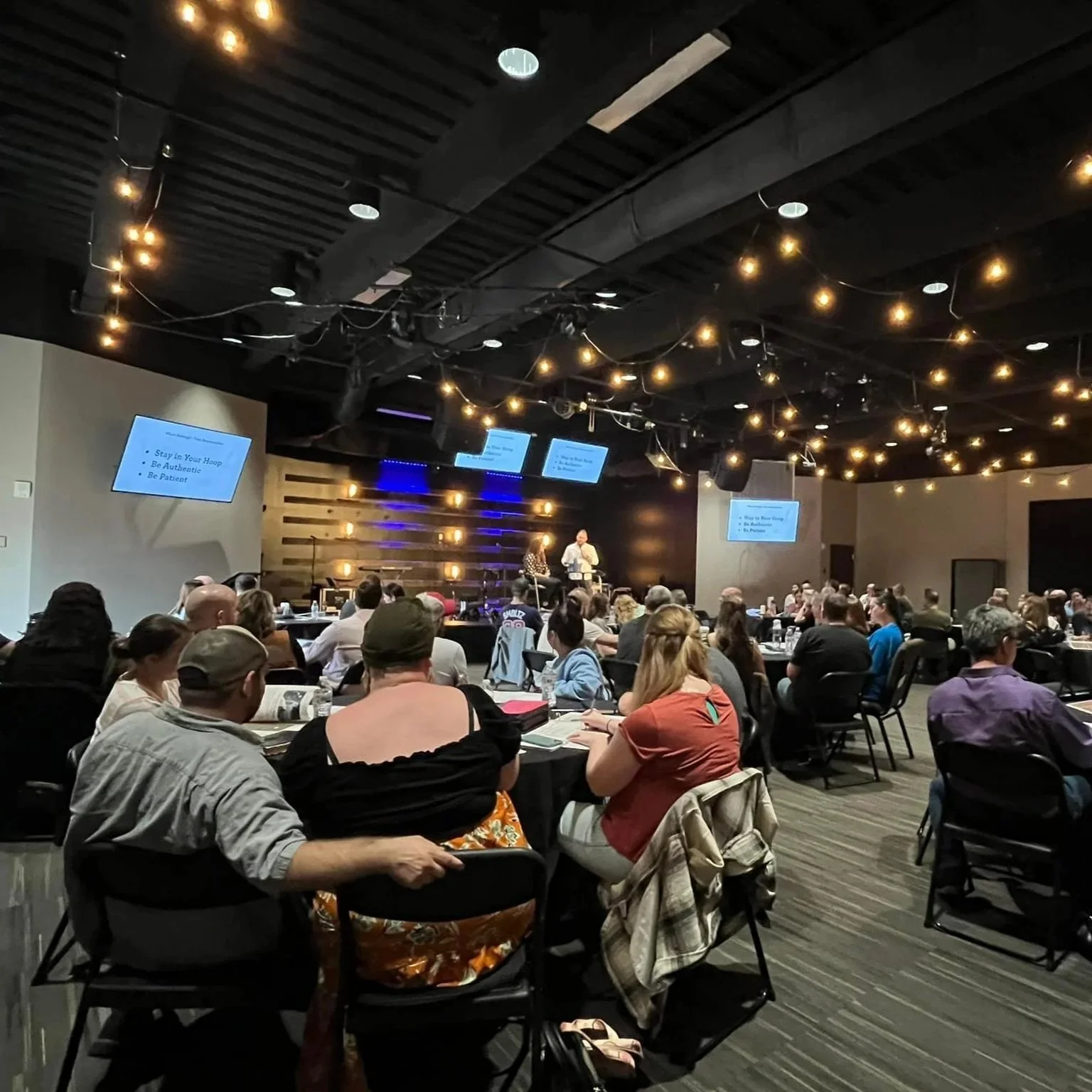 A large conference room filled with people sitting at round tables, watching a presentation on stage. The stage has two speakers, and three screens display the presentation slides. String lights are hanging from the ceiling, creating a warm atmosphere.