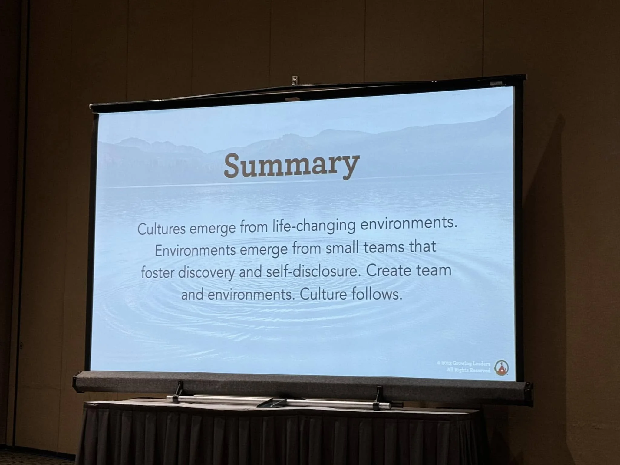 A presentation slide titled 'Summary' with a background of a lake and mountains. The slide contains text about cultures and environments, and a small logo at the bottom right corner.