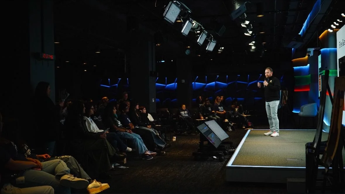 A speaker on a stage giving a presentation to a seated audience in a dark auditorium with blue lighting.