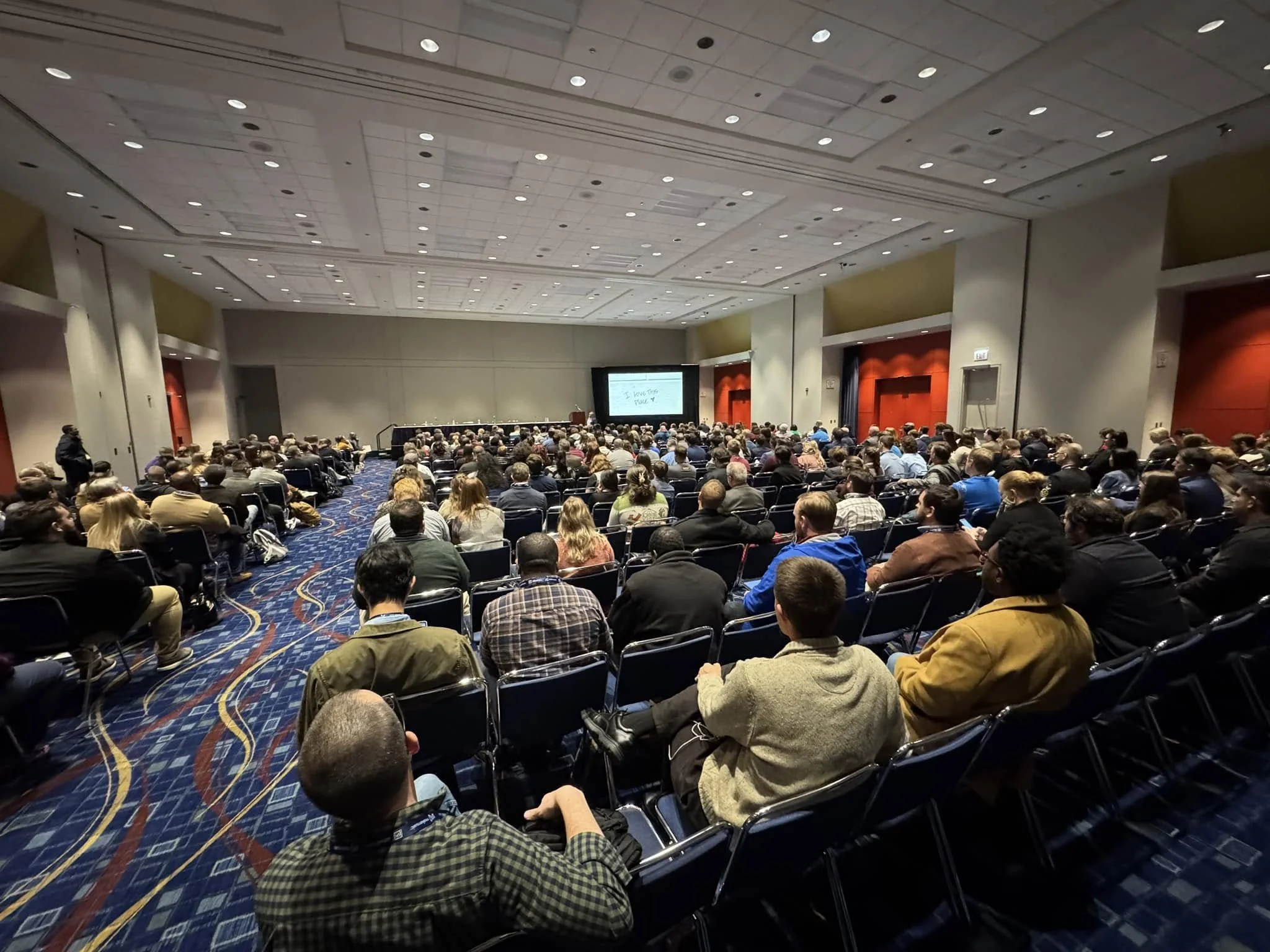 Large conference room filled with people seated, facing a screen at the front during a presentation.