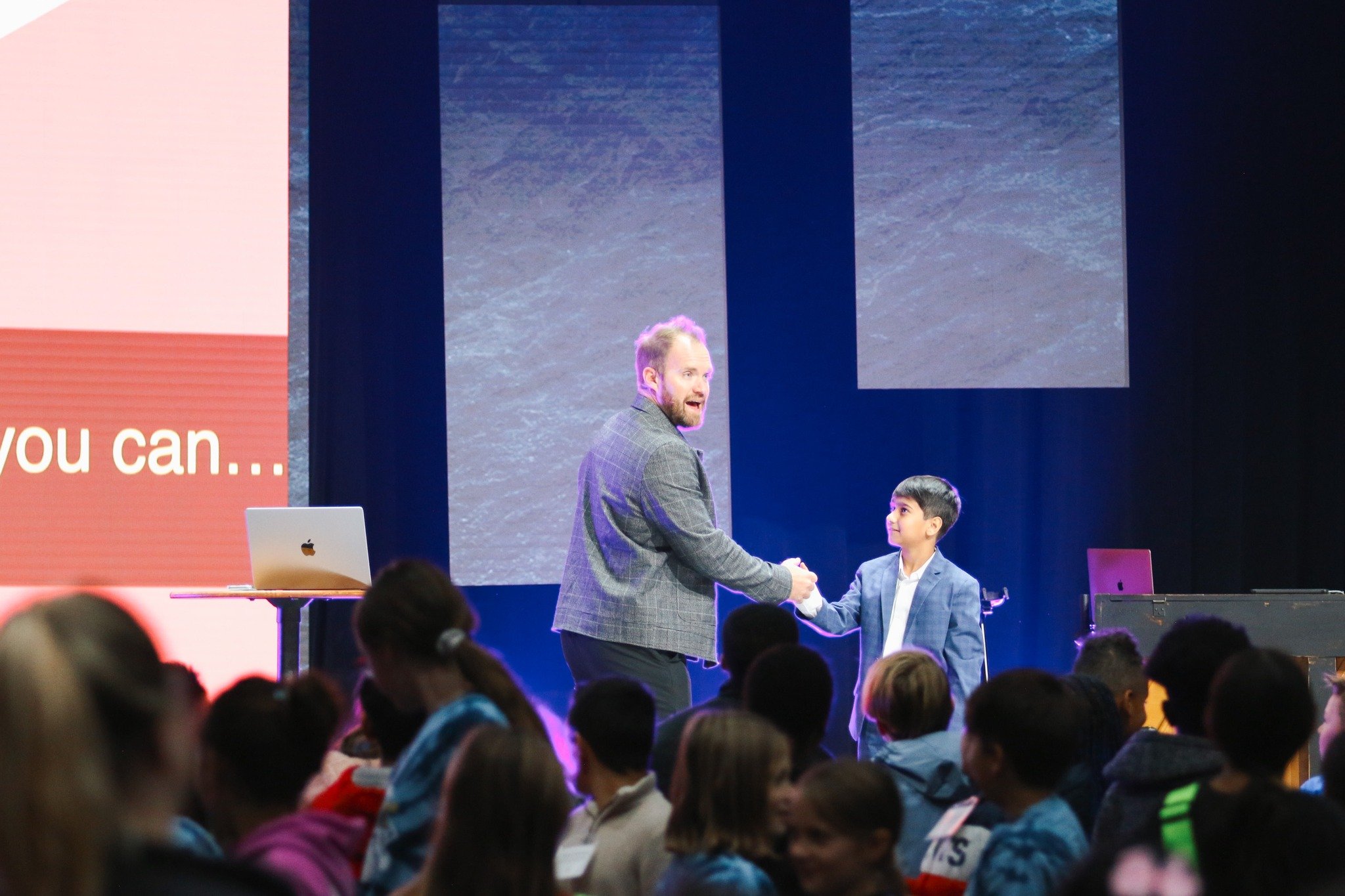 A man in a gray blazer shaking hands with a young boy in a blue blazer on stage during a presentation or event, with a large audience watching.