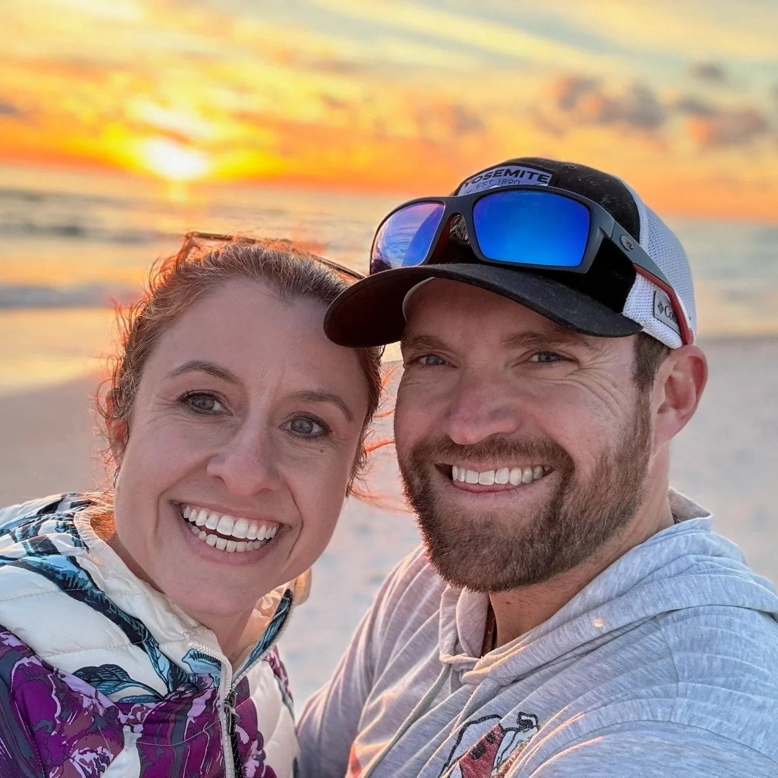 A smiling couple taking a selfie on the beach at sunset, with the ocean and colorful sky in the background.