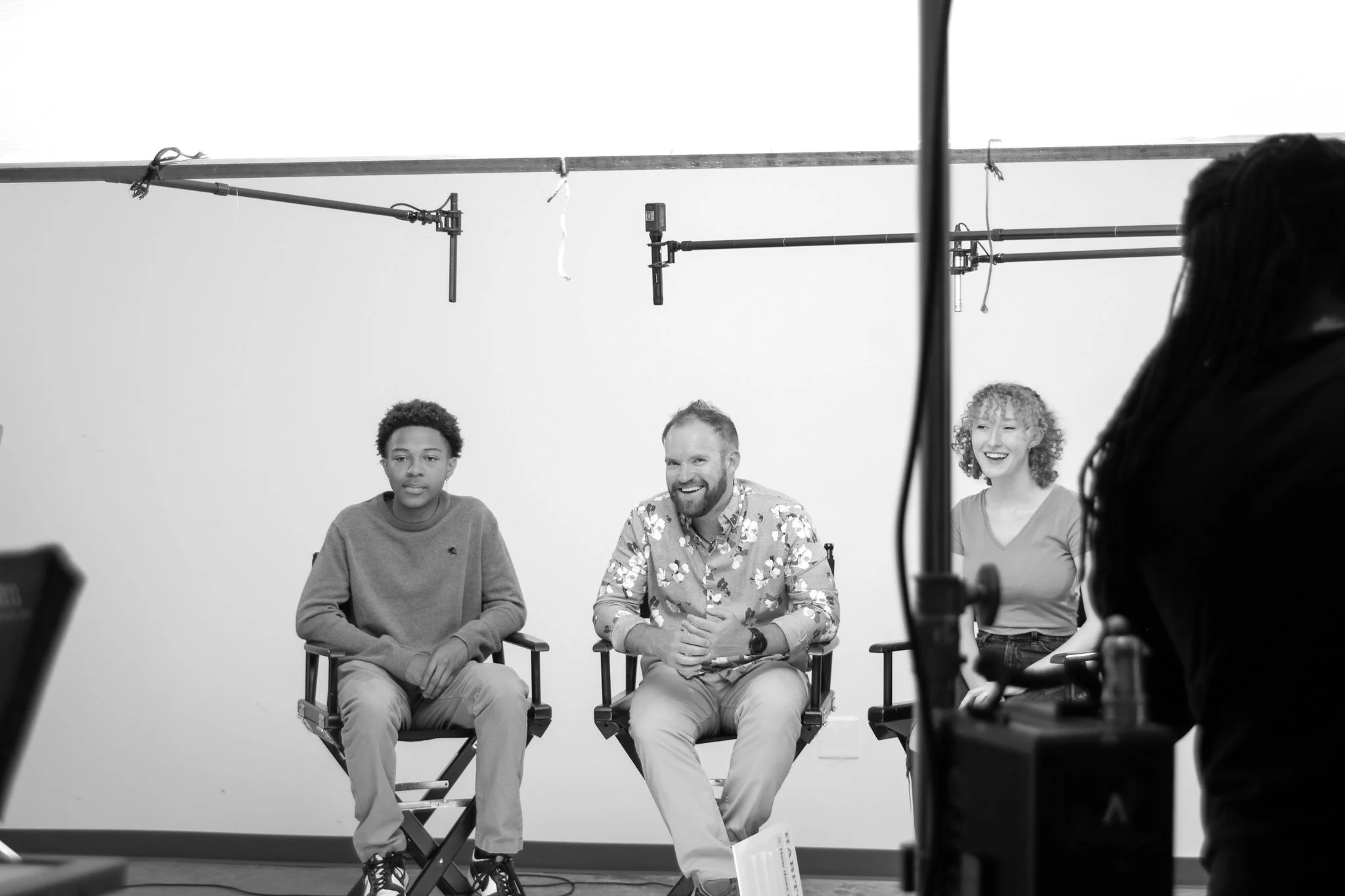 Three people sitting on director's chairs in a photography studio with a white background, two women and one man, with filming and lighting equipment partially visible.