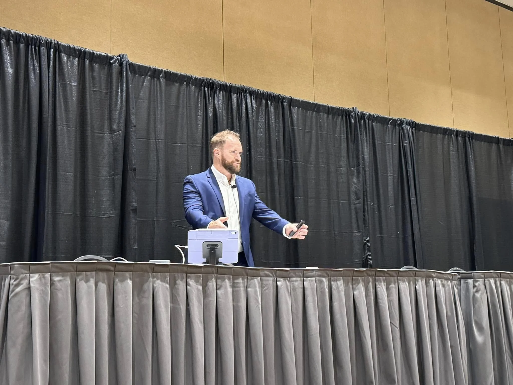 A man in a blue suit and white shirt stands behind a conference table with a black curtain backdrop, holding a microphone.