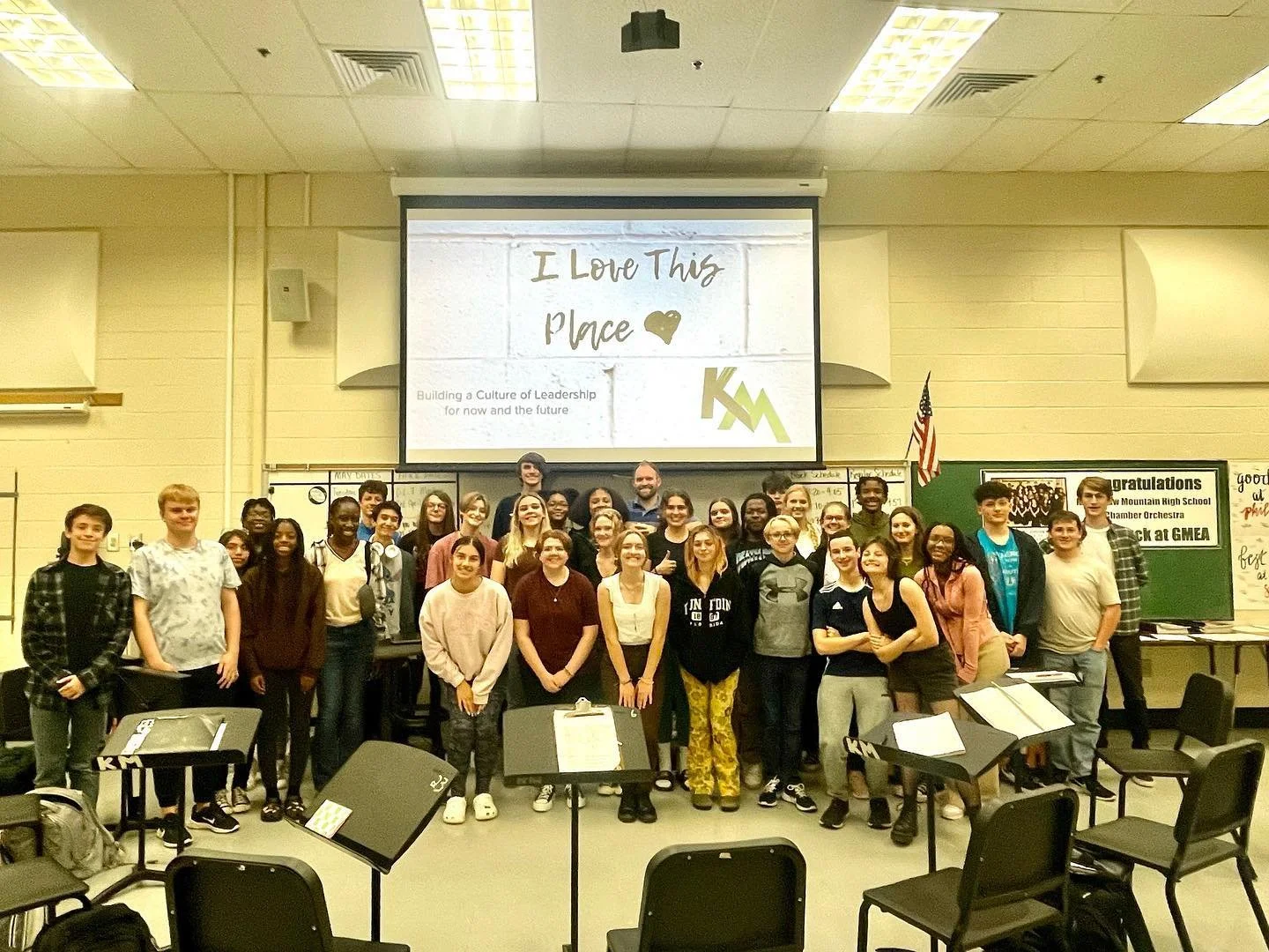 Group of students and teachers in a classroom posing for a photo, with a projection screen behind them displaying the message 'I Love This Place' and a logo.