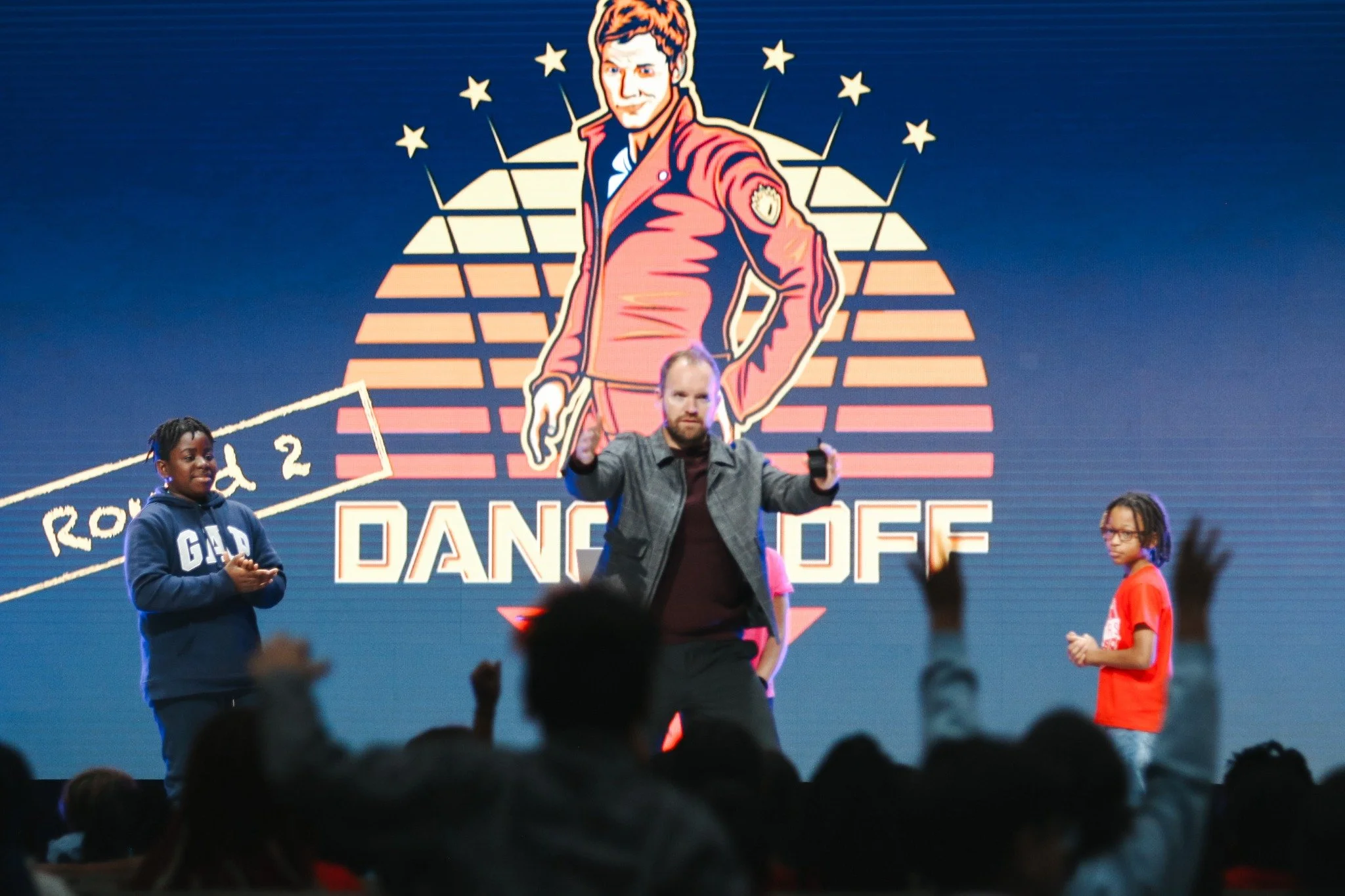 A man on stage with children around him, in front of a large graphic backdrop with a stylized image of a person in a red jacket. The backdrop features the words 'DANCE OFF' and stars. The audience is visible with some people raising their hands.
