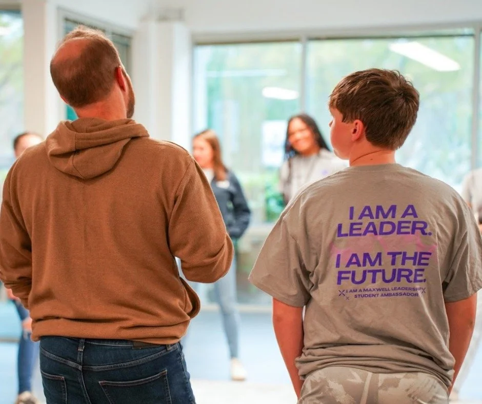 Young boy wearing a gray t-shirt with purple text that reads 'I AM A LEADER. I AM THE FUTURE.' standing in a room with large windows, listening to an adult male in a brown hoodie speaking, with several people in the background.