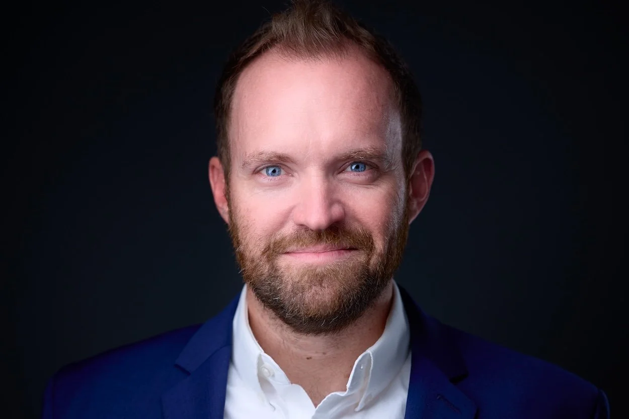 Headshot of a man with short brown hair, a beard, and blue eyes, wearing a white shirt and navy blazer against a dark background.