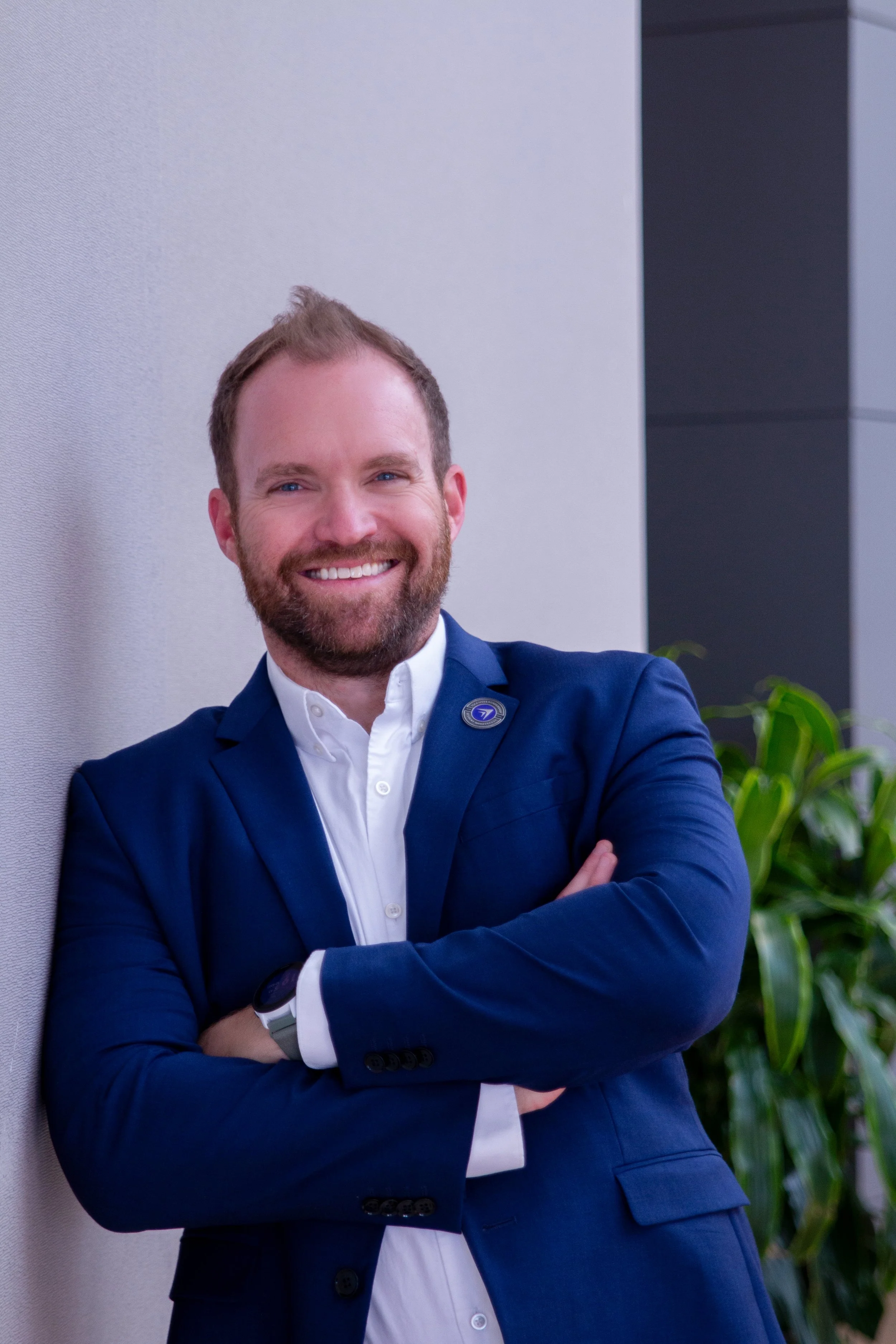 Smiling man in a blue suit and white shirt standing with arms crossed next to a gray wall and green plant.