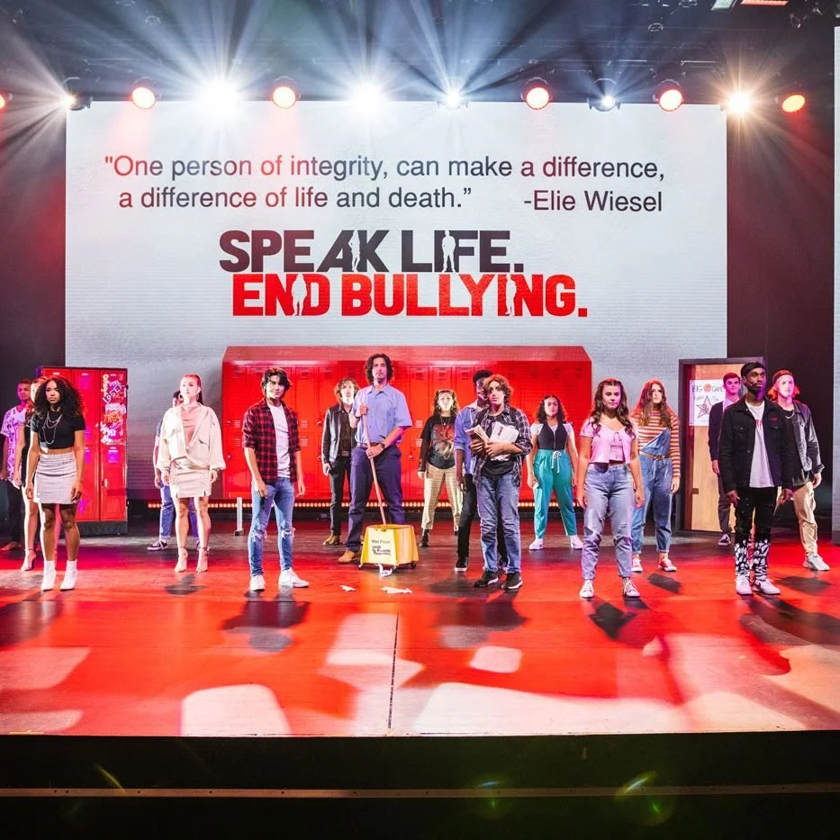 Group of teenagers standing on stage during anti-bullying presentation with a large screen behind them displaying the message 'Speak Life. End Bullying.' and a quote by Elie Wiesel.