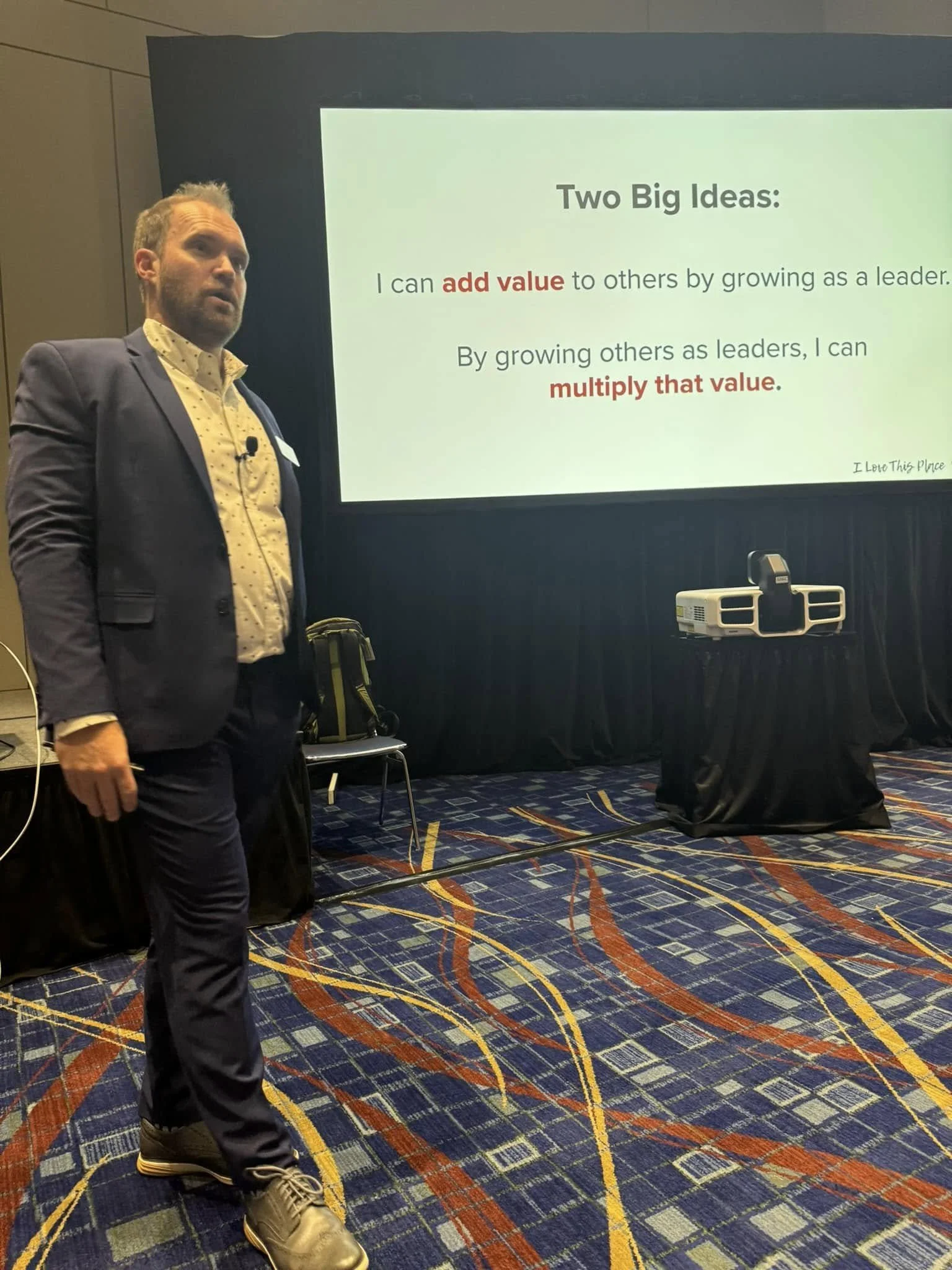 A man in a suit standing in front of a presentation slide that reads 'Two Big Ideas: I can add value to others by growing as a leader. By growing others as leaders, I can multiply that value.'