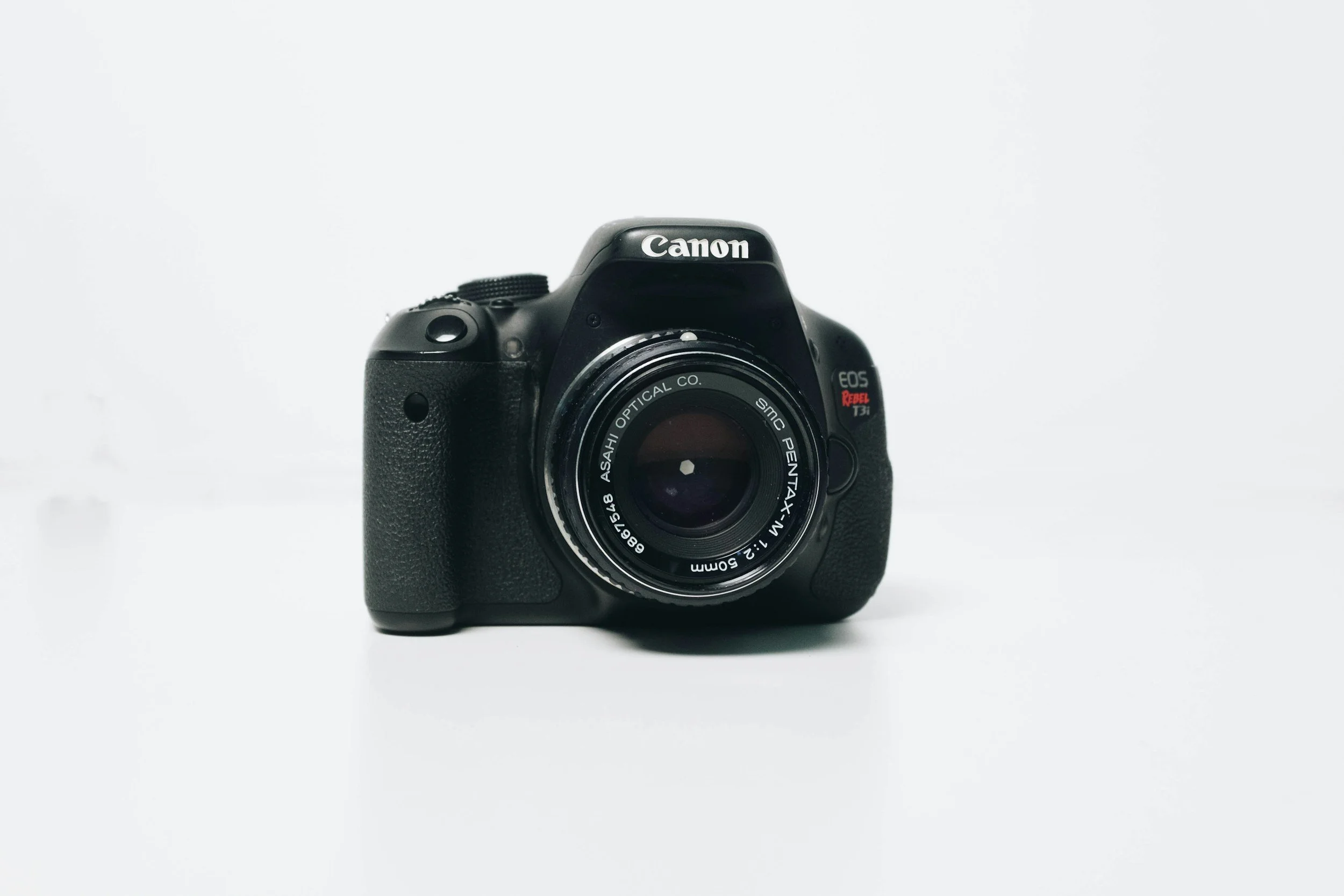 A black Canon EOS Rebel T3i camera with a lens attached, placed on a plain white surface and background.