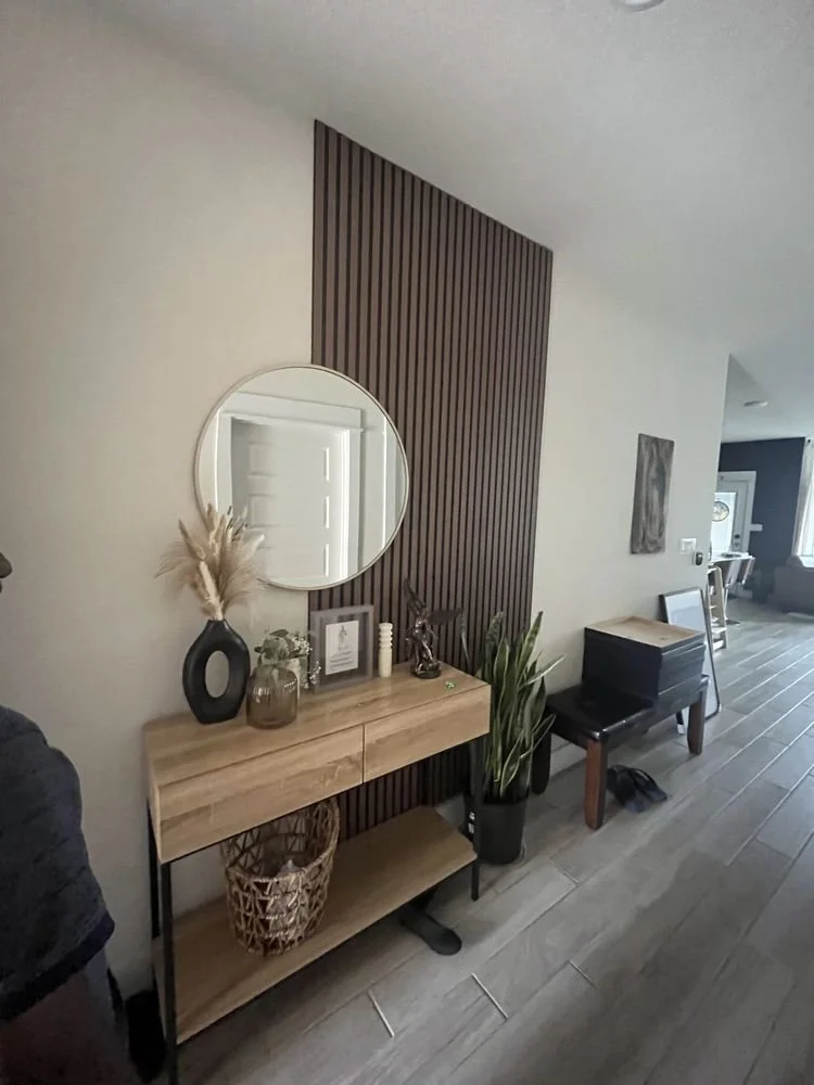 Interior view of a modern home entryway with a wooden console table, decorative items including vases and frames, a mirror on the wall, a tall potted plant, and a bench. The space features neutral wall colors, wood accents, and tiled flooring.