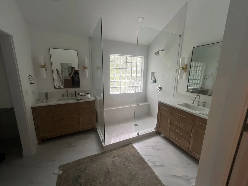Modern bathroom with dual vanities, wooden cabinets, large mirror, glass shower enclosure, tiled shower area, glass block window, marble flooring, and soft white lighting.