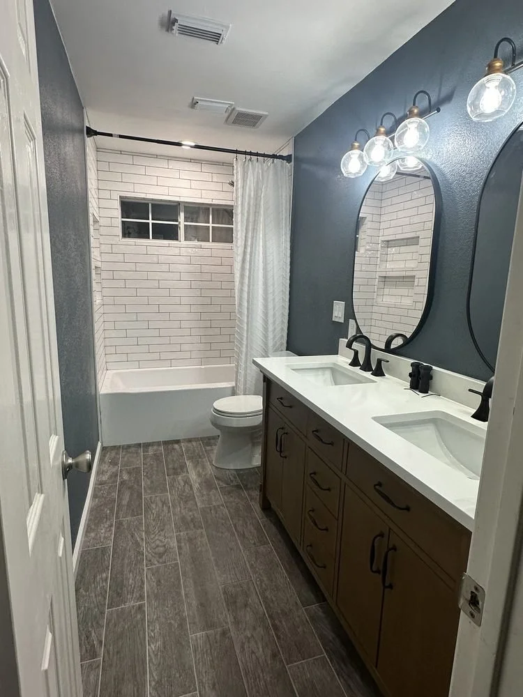 A modern bathroom with a double sink vanity, two mirrors, and three light fixtures above the mirrors. The vanity has dark wooden cabinets. At the back, there is a bathtub with a white subway tile surround and a small window, partially covered by a cu