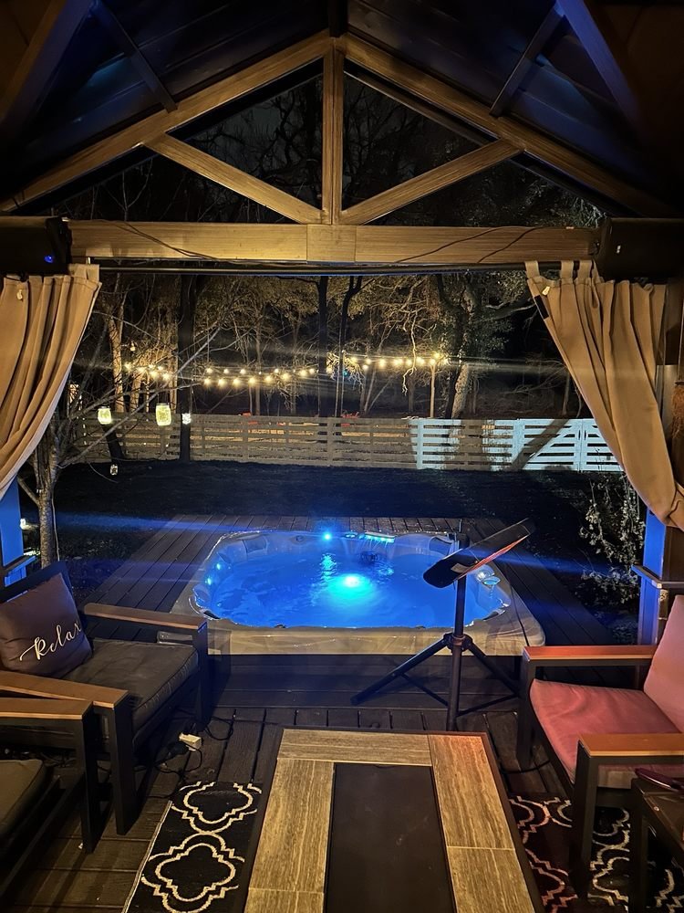 Cozy outdoor patio with a hot tub illuminated with blue lights, surrounded by string lights, with outdoor furniture including chairs and a rug, at night.